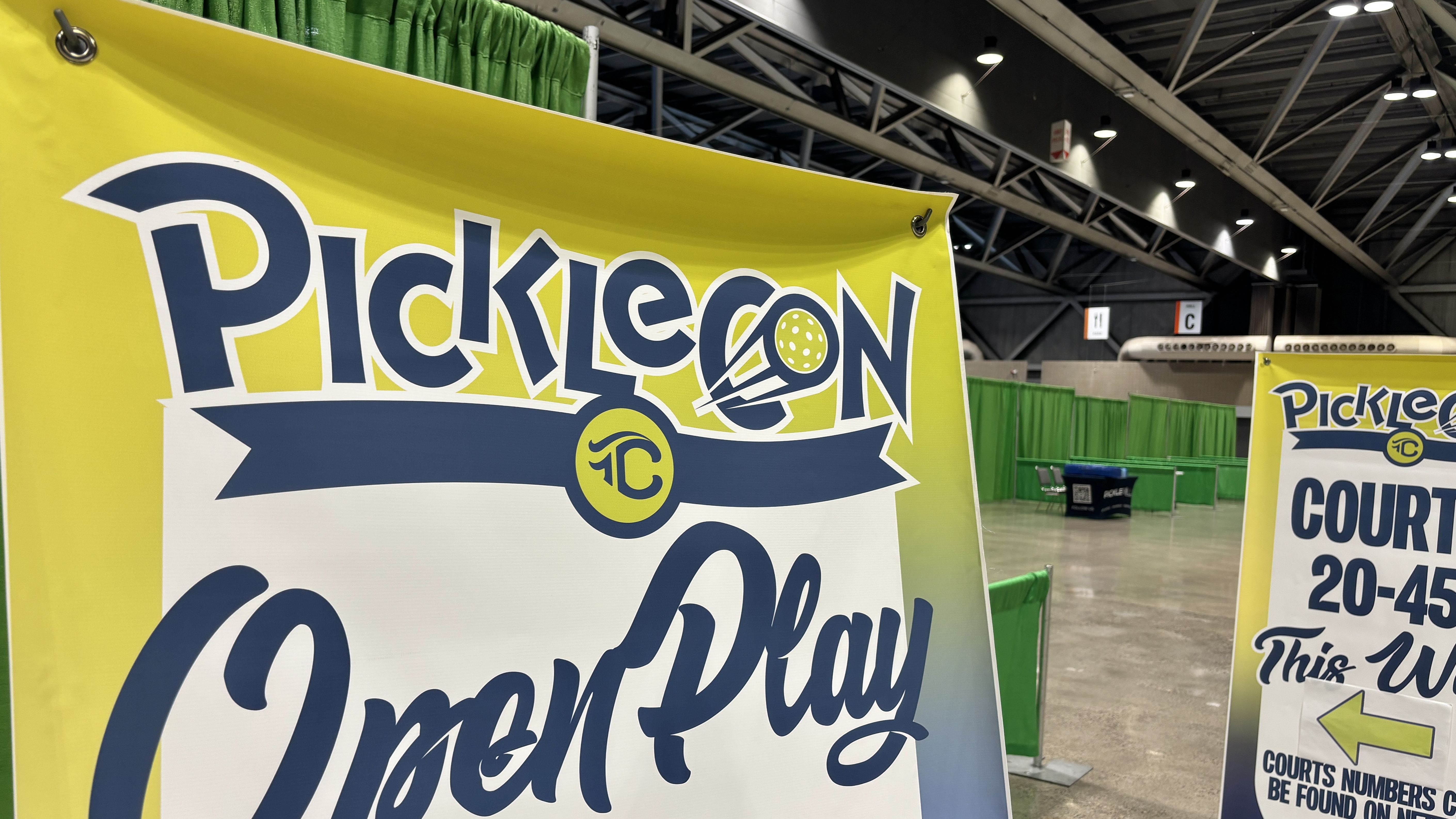 Bright yellow banner with bold blue and white lettering and a pickleball icon, displayed in a large convention hall with green partition walls and exposed ceiling beams.