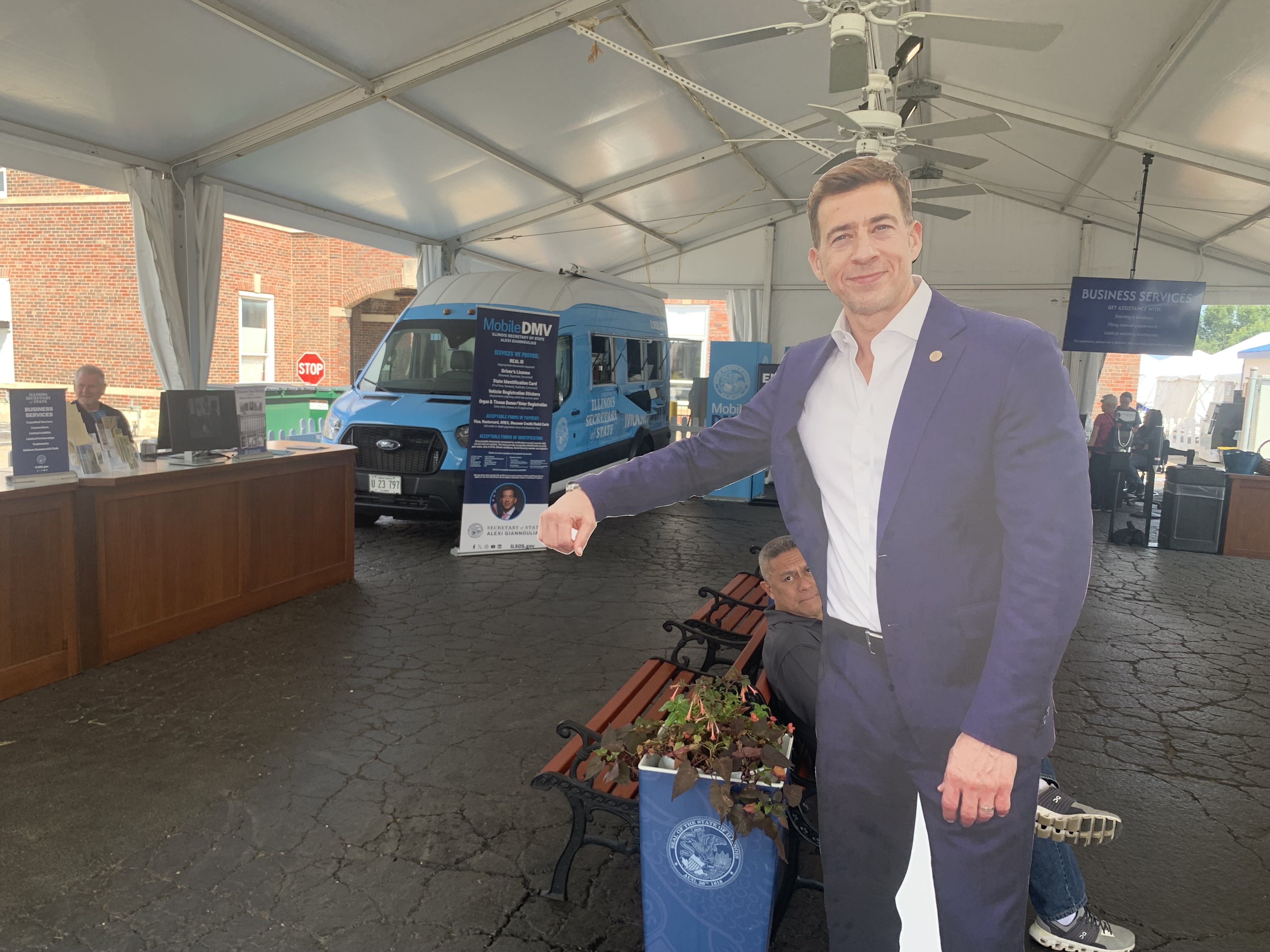 Inside a white event tent, a life-size cardboard cutout of a smiling man in a blue suit stands beside a blue Mobile DMV van; tables, brochures, and a "BUSINESS SERVICES" sign are in the background.