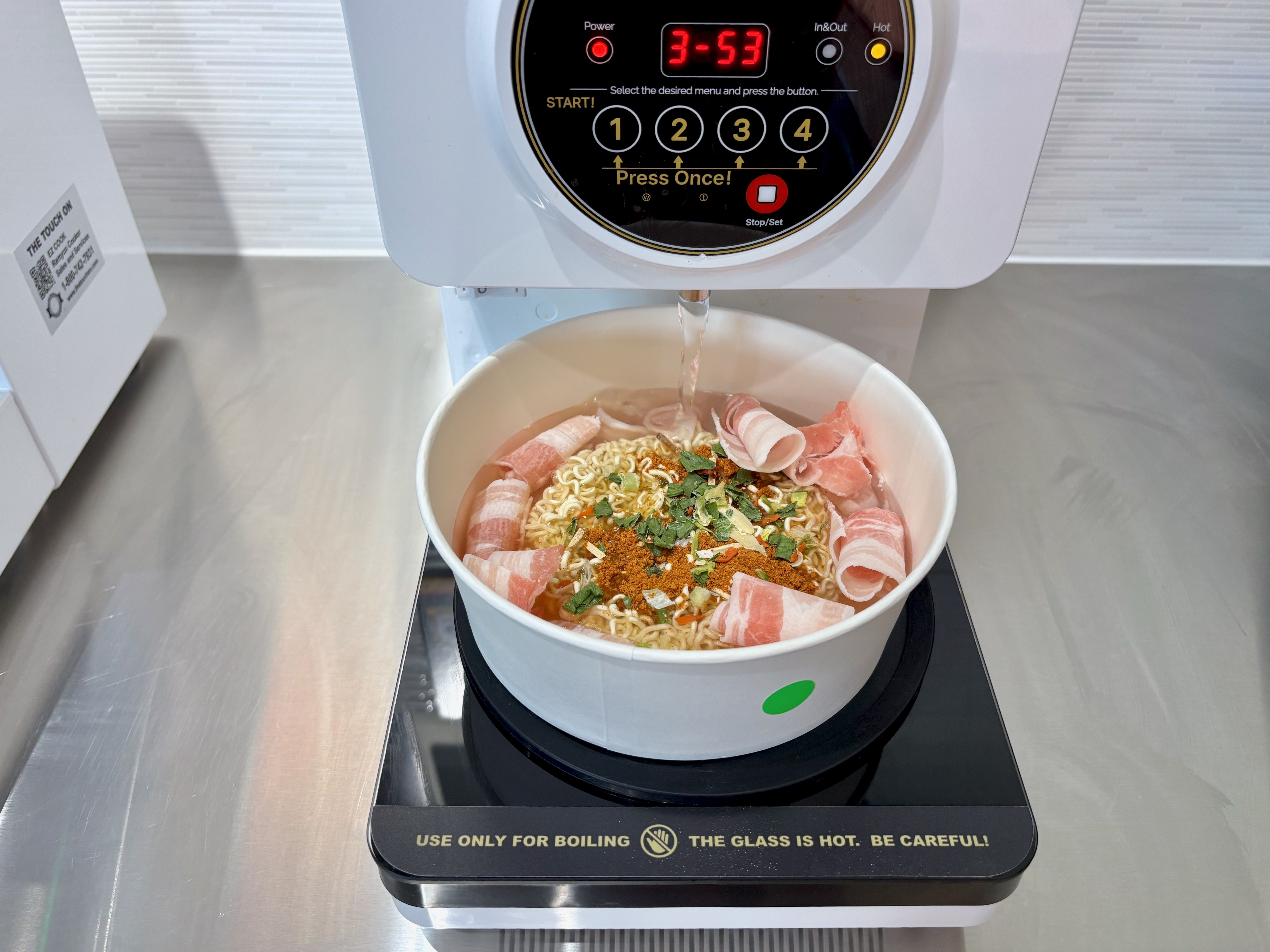 White noodle-boiling machine with a circular black control panel and red digital display; hot water flows into a bowl of ramen topped with rolled ham slices, greens, and spices on a stainless counter.