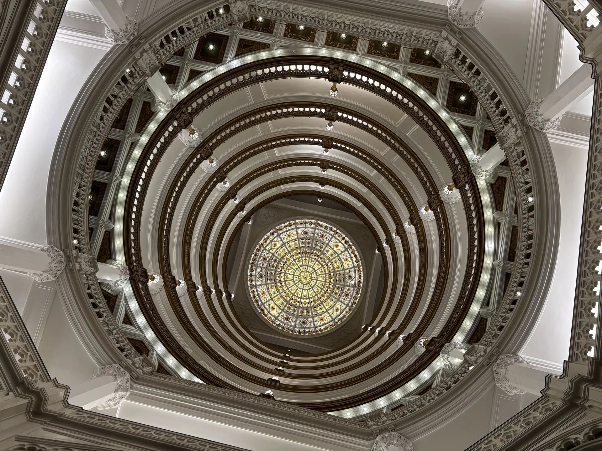 View looking up at a spiral staircase with white and brown railings, circular lights, and a decorative stained-glass dome ceiling in shades of yellow and brown.