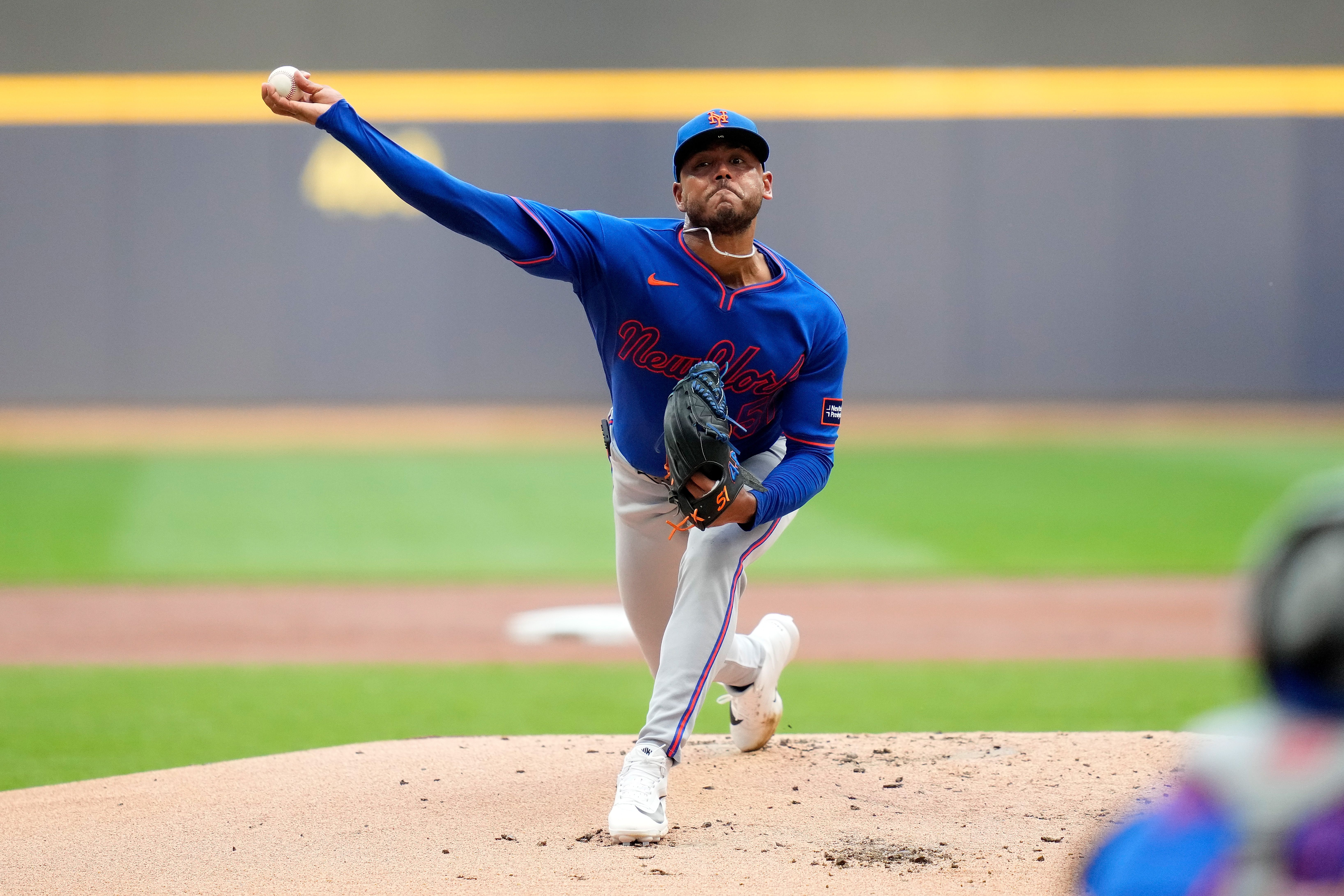 Baseball pitcher in a blue New York Mets uniform throws a pitch from the mound, wearing a blue cap and glove, with a blurred gray wall and green field behind.
