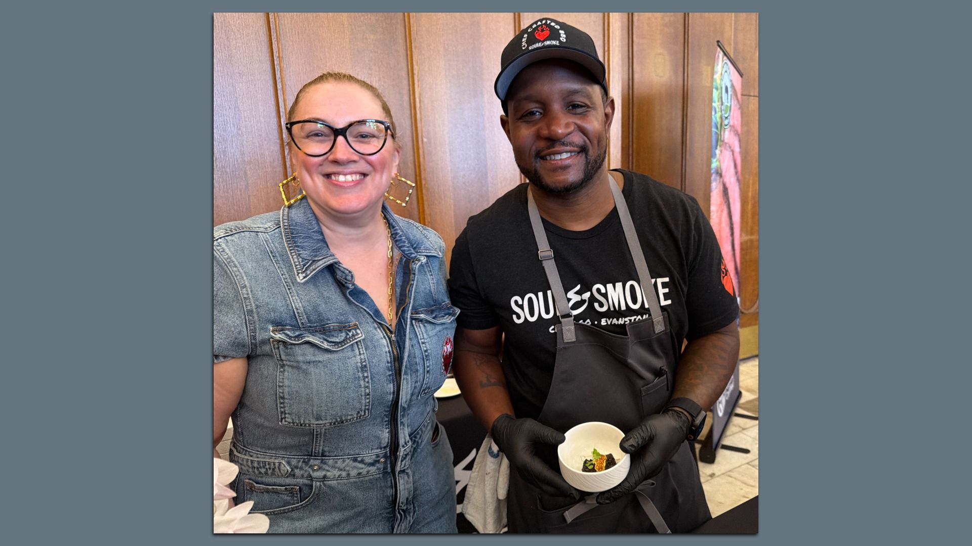 Two smiling people pose indoors in front of wooden wall panels. Left: a woman with glasses and a denim shirt. Right: a man in a black shirt, cap and apron, holding a cup of food.