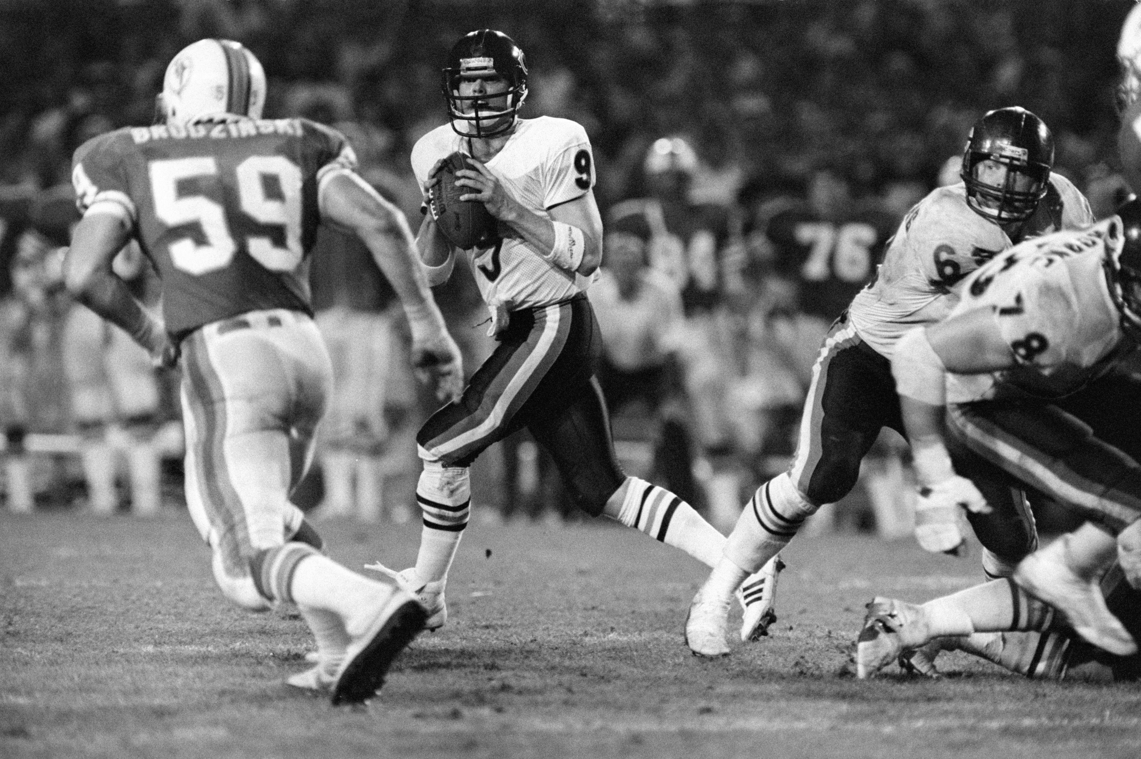 Black-and-white photo of a football quarterback in white jersey #9 holding the ball, preparing to throw, as opponents in dark helmets and white pants advance during a game under stadium lights.