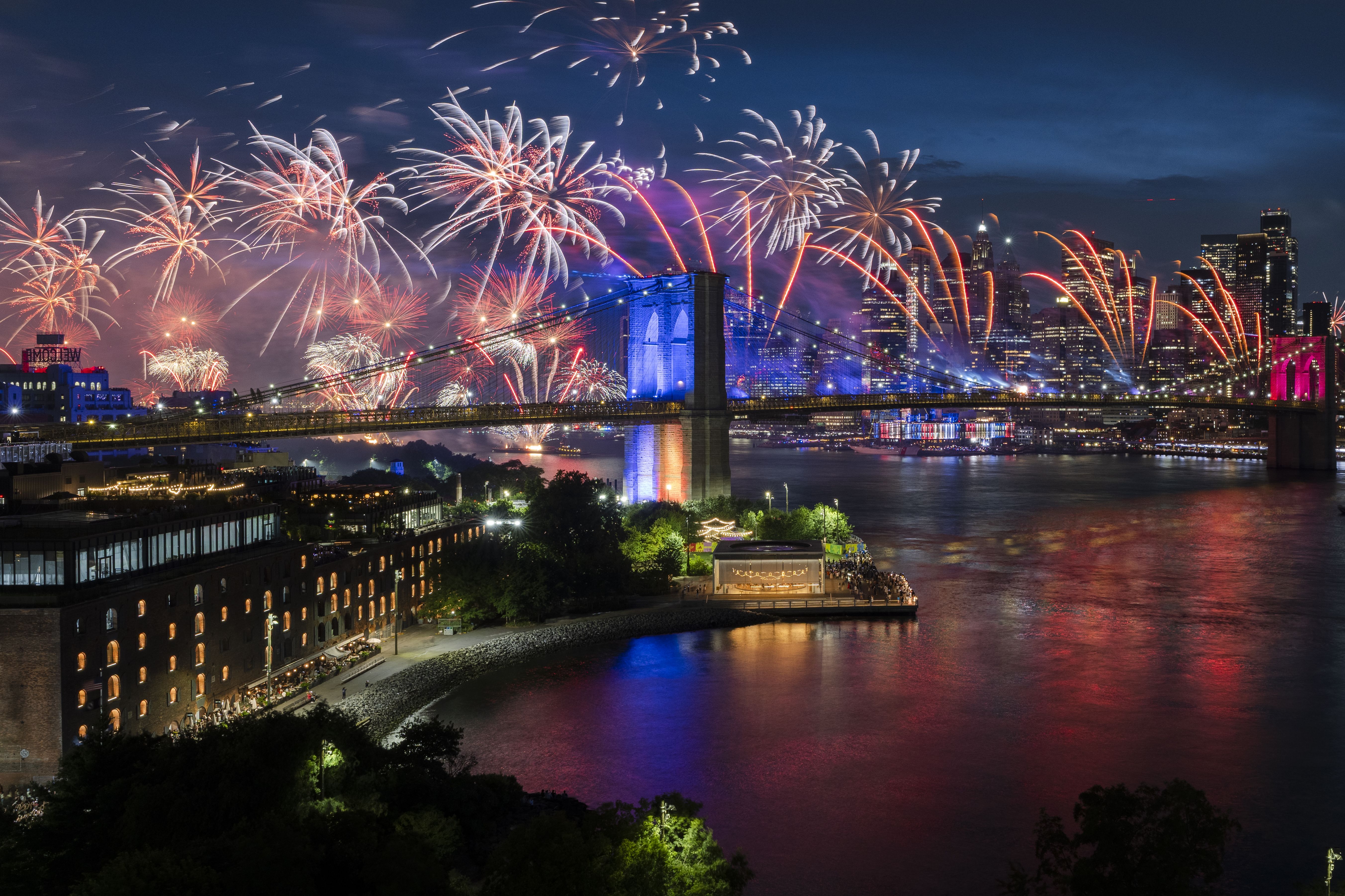 Fireworks burst above the Brooklyn Bridge and Lower Manhattan skyline during the Macy's Fourth of July Fireworks show, with colorful reflections shimmering across the East River in New York on July 4, 2026.