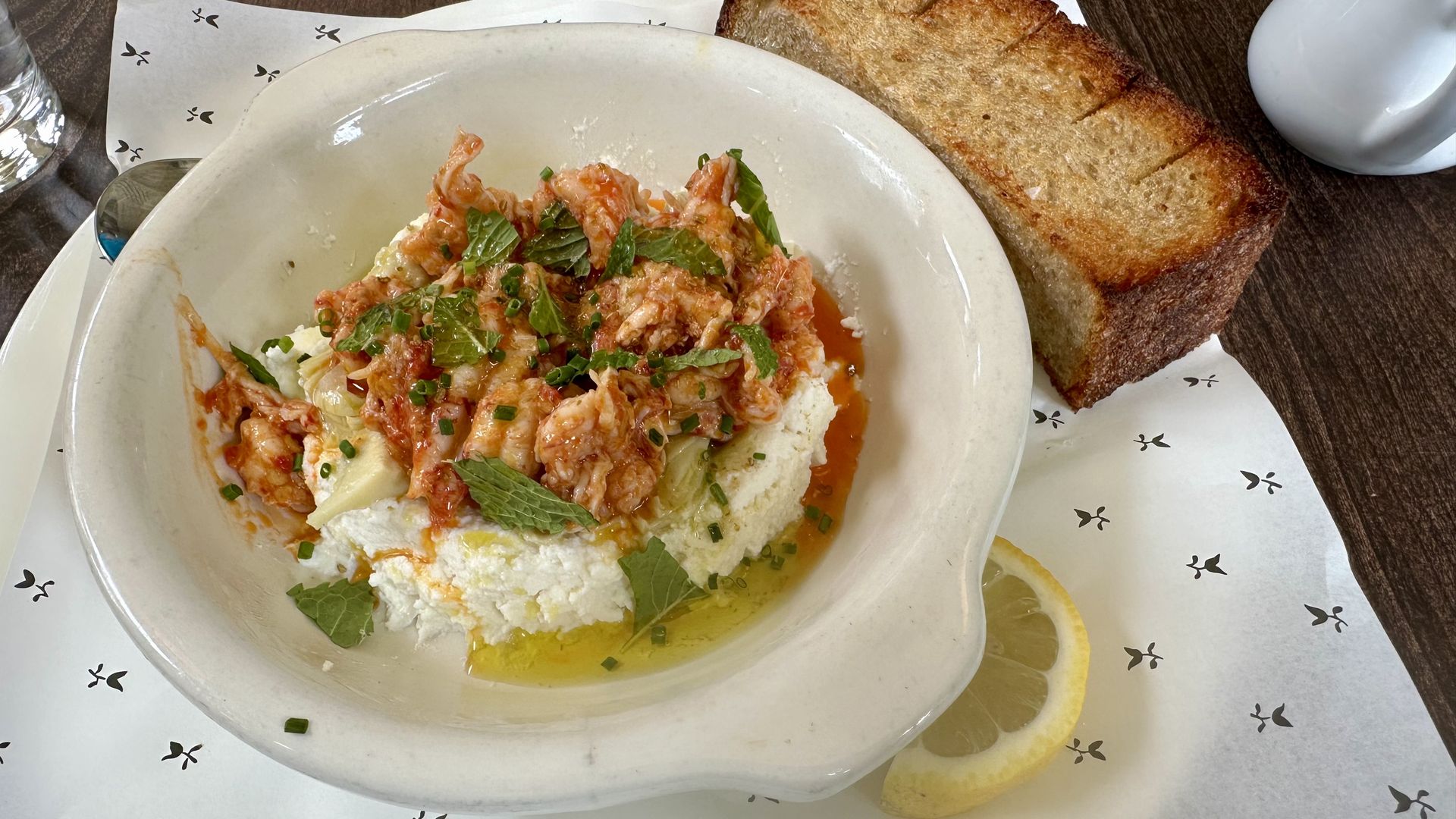 Top-down view of a white bowl filled with ricotta topped with crawfish, garnished with mint and chives; lemon wedges and toasted bread on a dark wooden table.