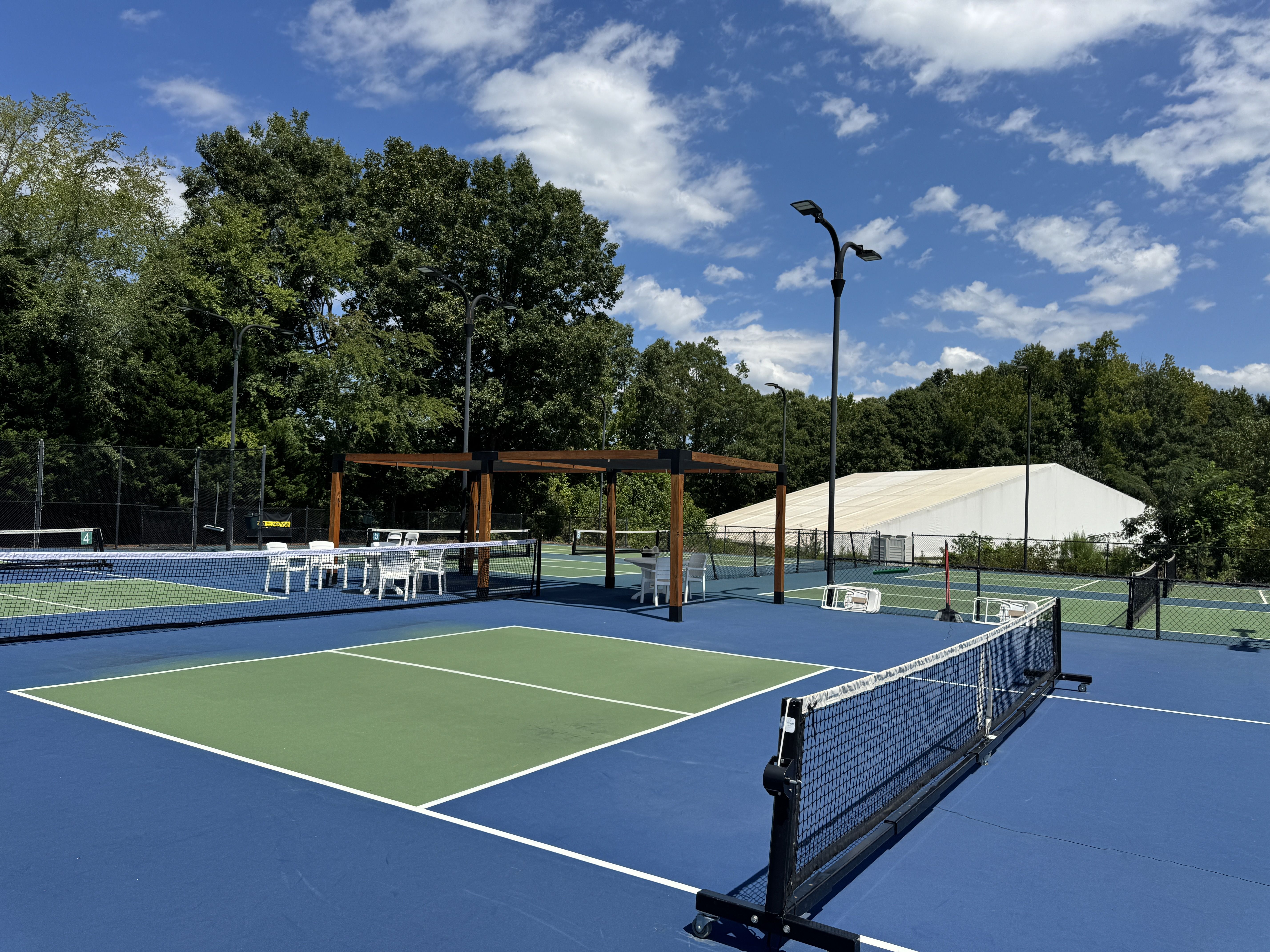 Outdoor pickleball courts with blue and green surfaces, surrounded by trees and a white tent. White chairs and tables are placed under a wooden pergola and near the courts under sunny sky.