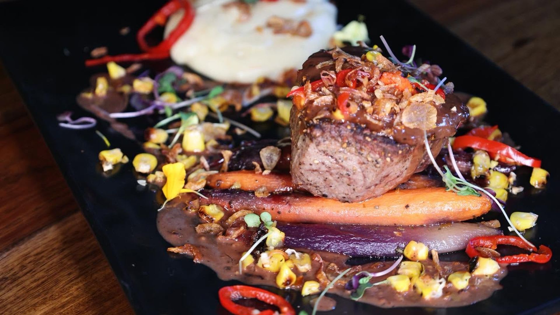 Filet mignon with an onion-pepper relish on a bed of roasted vegetables (carrot, corn, red onion, peppers), microgreens, and brown sauce on a black plate; mashed potatoes blur in the background.