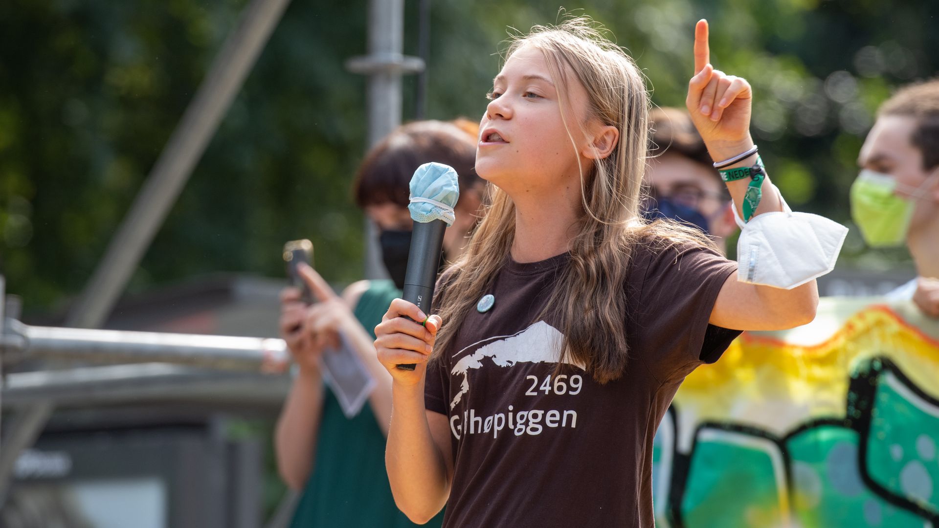 A teen girl speaks into a microphone on an outdoor platform, behind her people hold up a banner