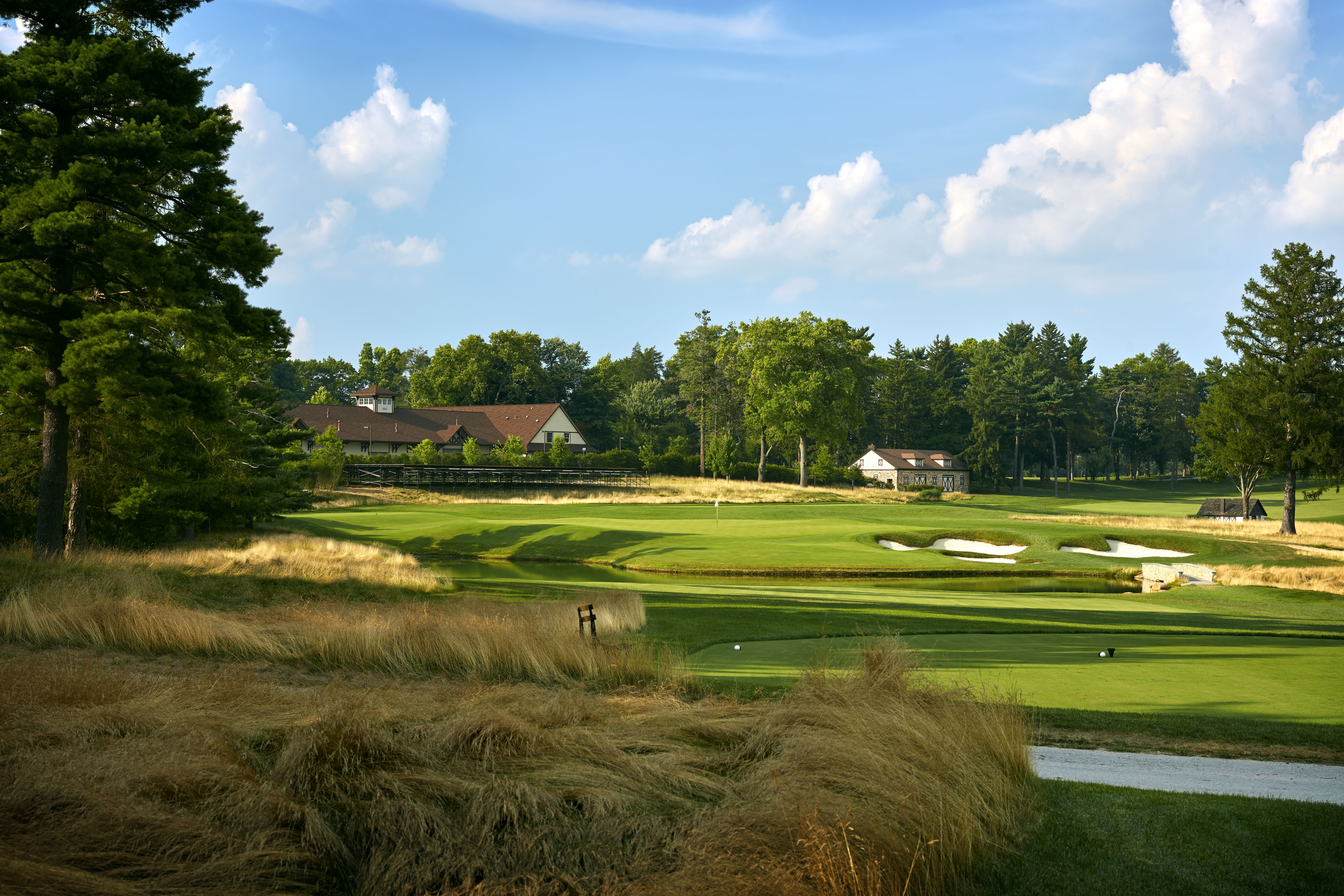 Sunlit golf course with green fairways, sand bunkers, and a small pond. A brown-roofed building sits among trees on the left, while tall pines and a blue sky with white clouds fill the scene.