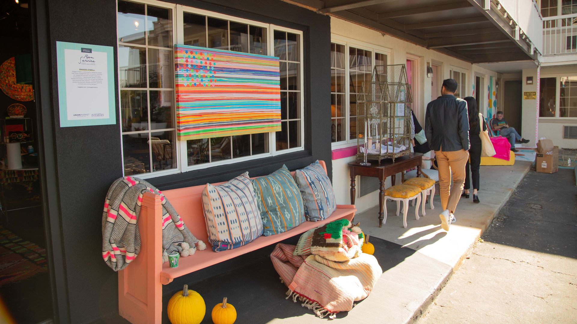 Outdoor corridor of motel or hotel with peach bench, colorful pillows and blankets, two yellow pumpkins, colorful abstract American flag painting, people walking away, and decorative table.