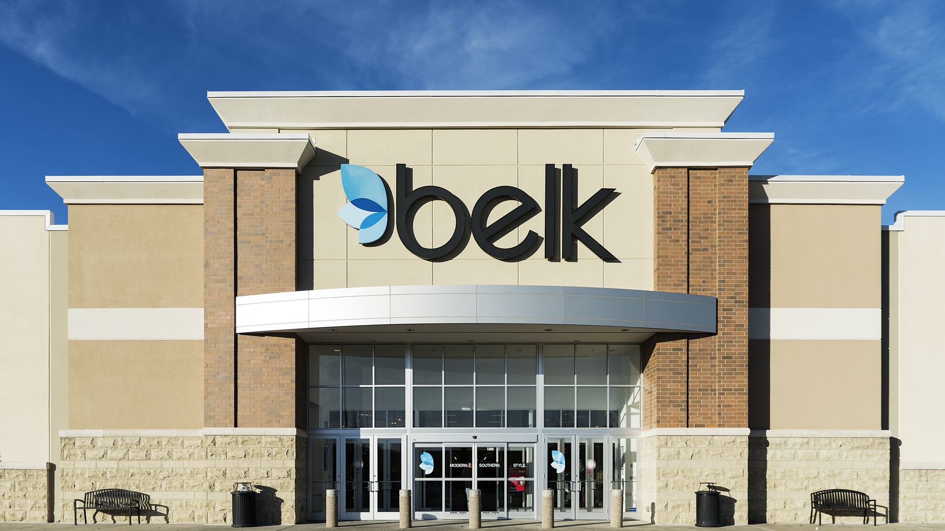 Front entrance of a Belk department store with beige and brick exterior under a blue sky, featuring large black and blue Belk logo above the glass doors.