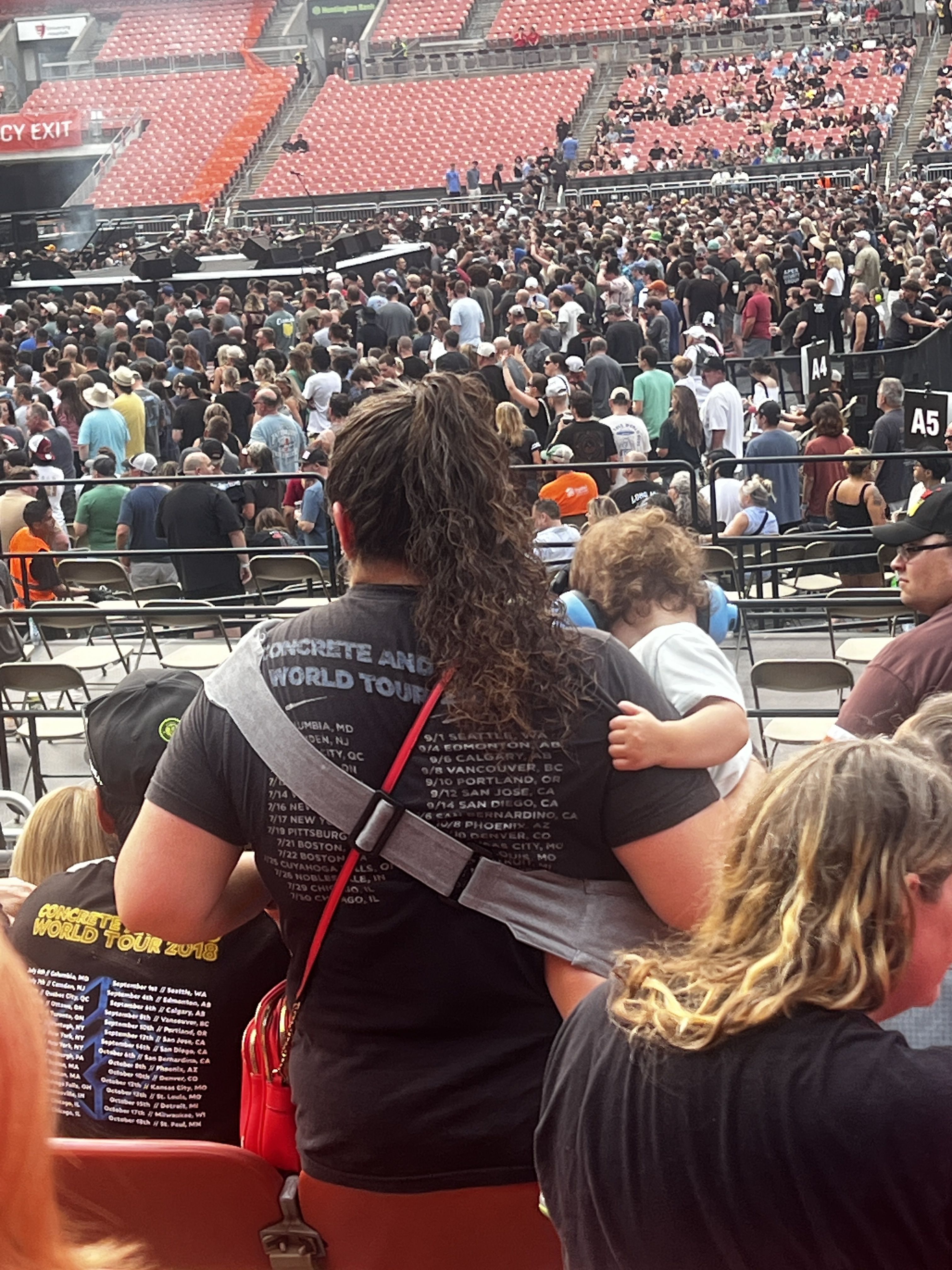 Large crowd at a stadium concert, stage left, people near barricades. Bright red seats in the background. Foreground shows a man with long hair wearing a dark T-shirt and a crossbody strap.
