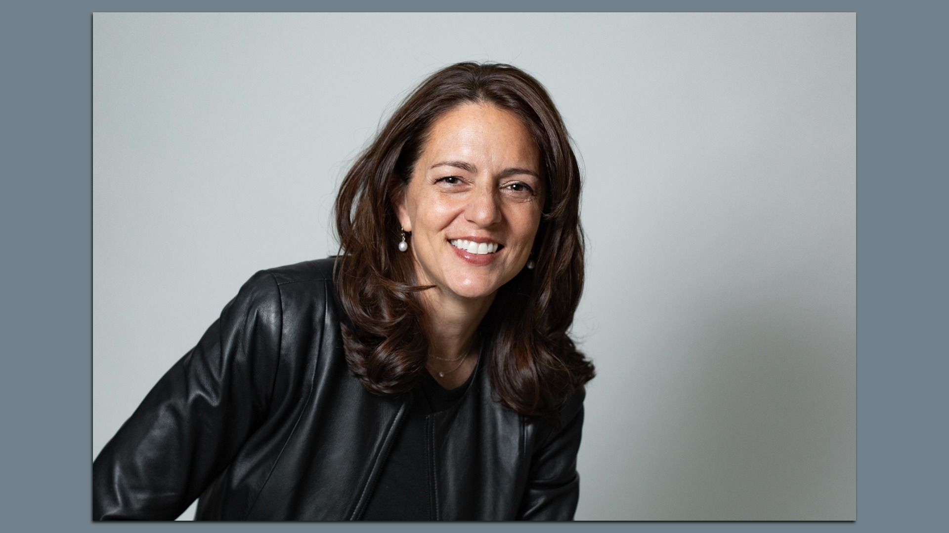 Portrait of a smiling woman with shoulder-length brown hair, wearing a black leather jacket and pearl earrings, against a light gray backdrop.