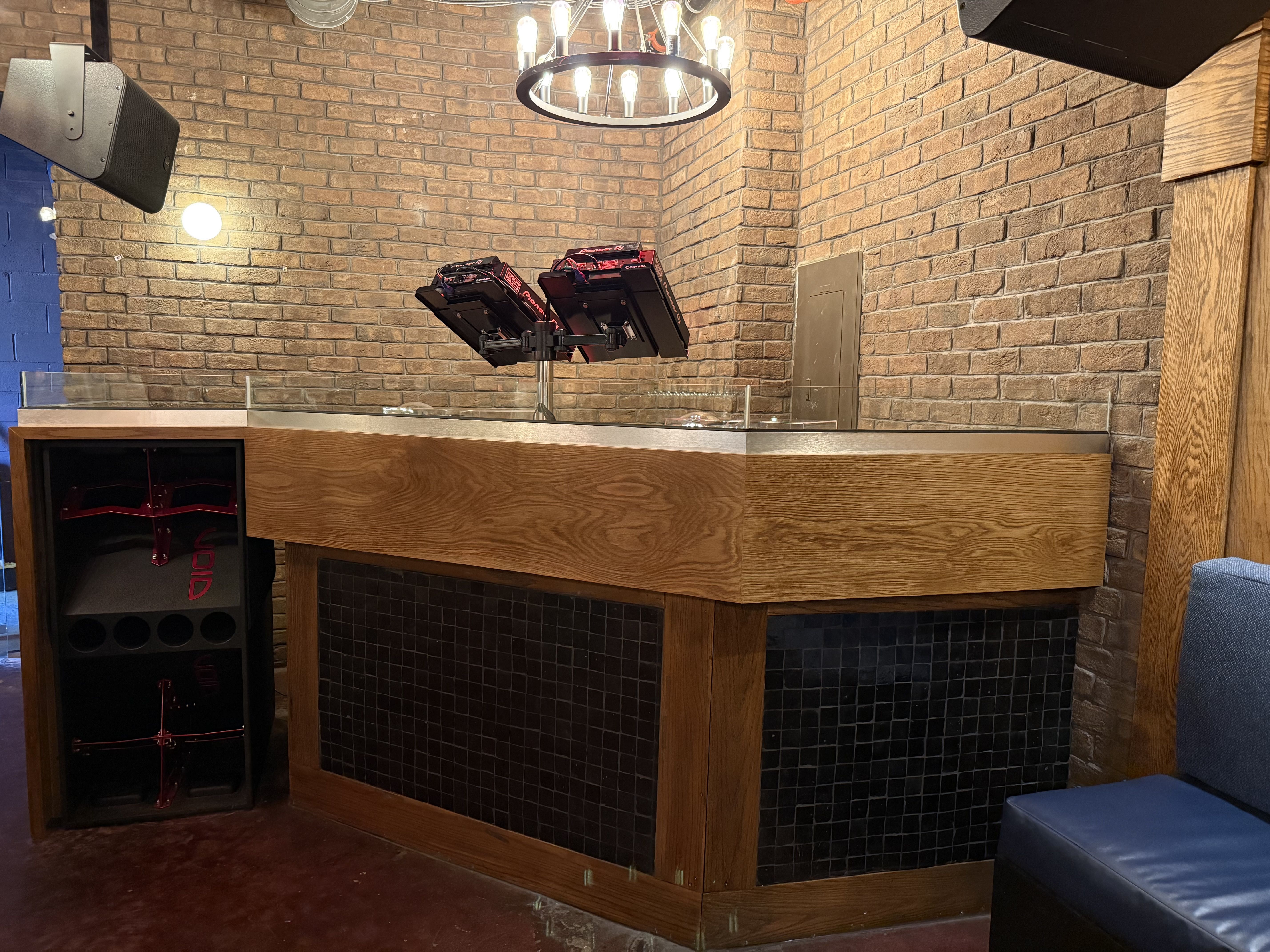 Dimly lit brick-walled interior with a circular chandelier, speakers mounted to the walls, a DJ stand with Pioneer gear, wooden counter and black-tiled base, blue wall on left.