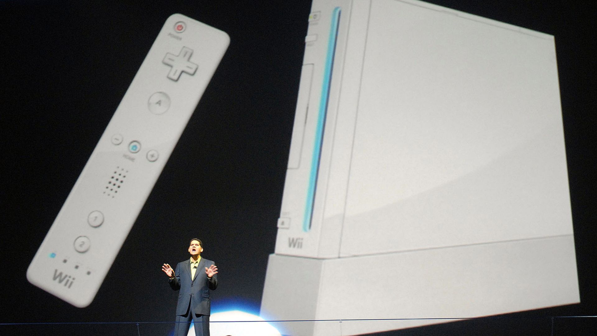Photo of a man on stage in front of a giant projection of the Nintendo Wii home console and controller