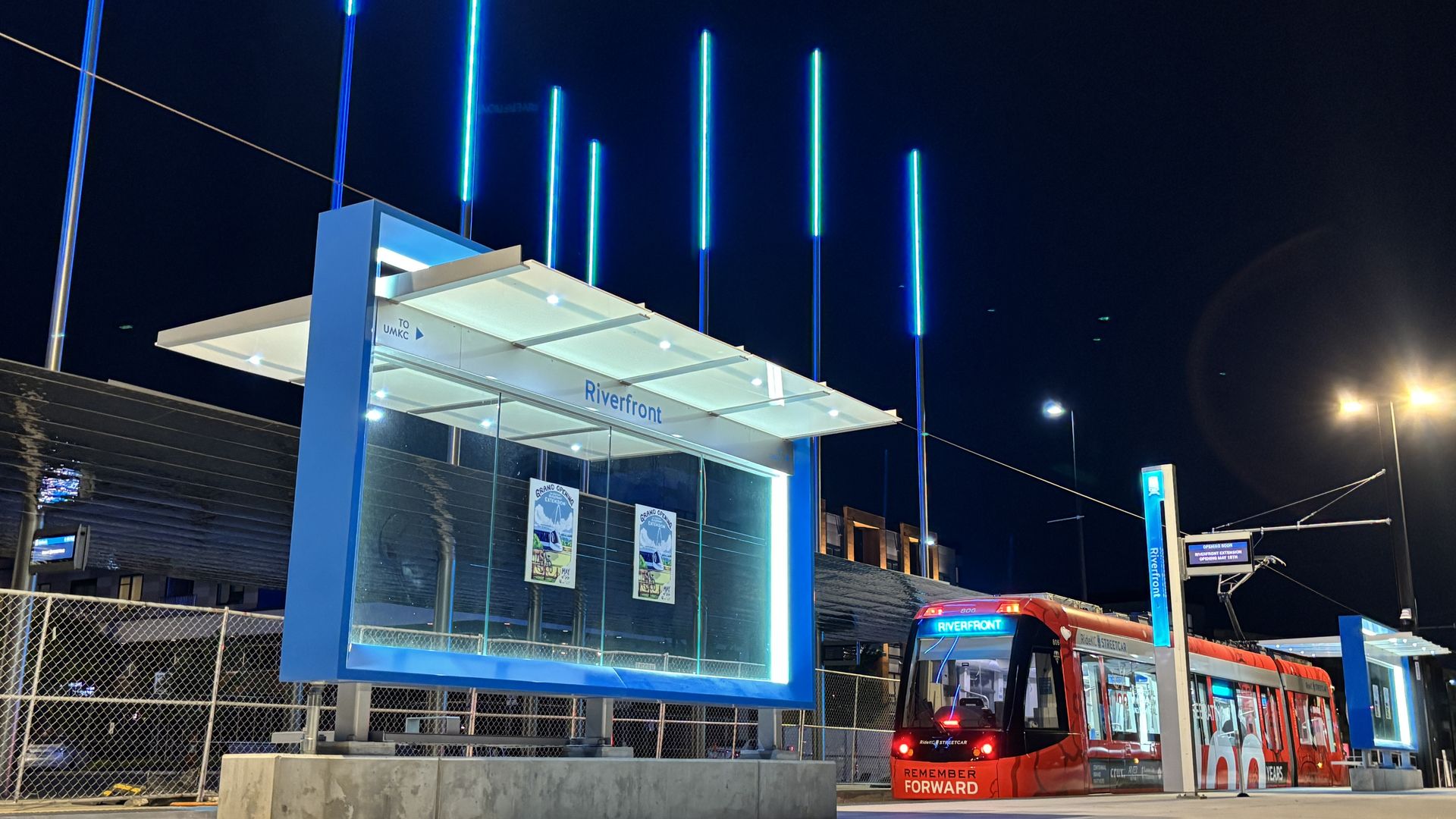 Night scene of a modern Riverfront transit entrance with glowing blue vertical light poles, a glass canopy, and a red bus at the curb; a chain-link fence is in the foreground.