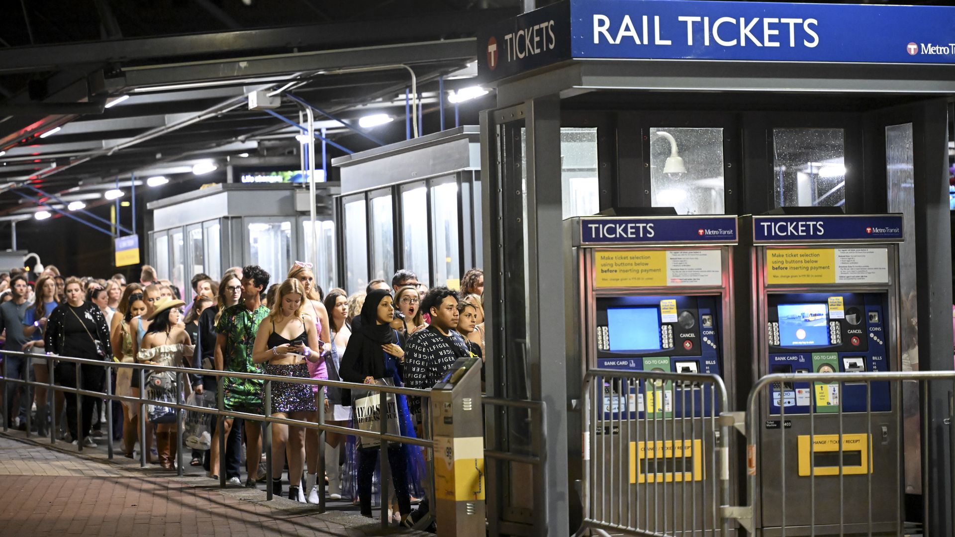 People wait in line at a transit station, with payment kiosks in front of them 