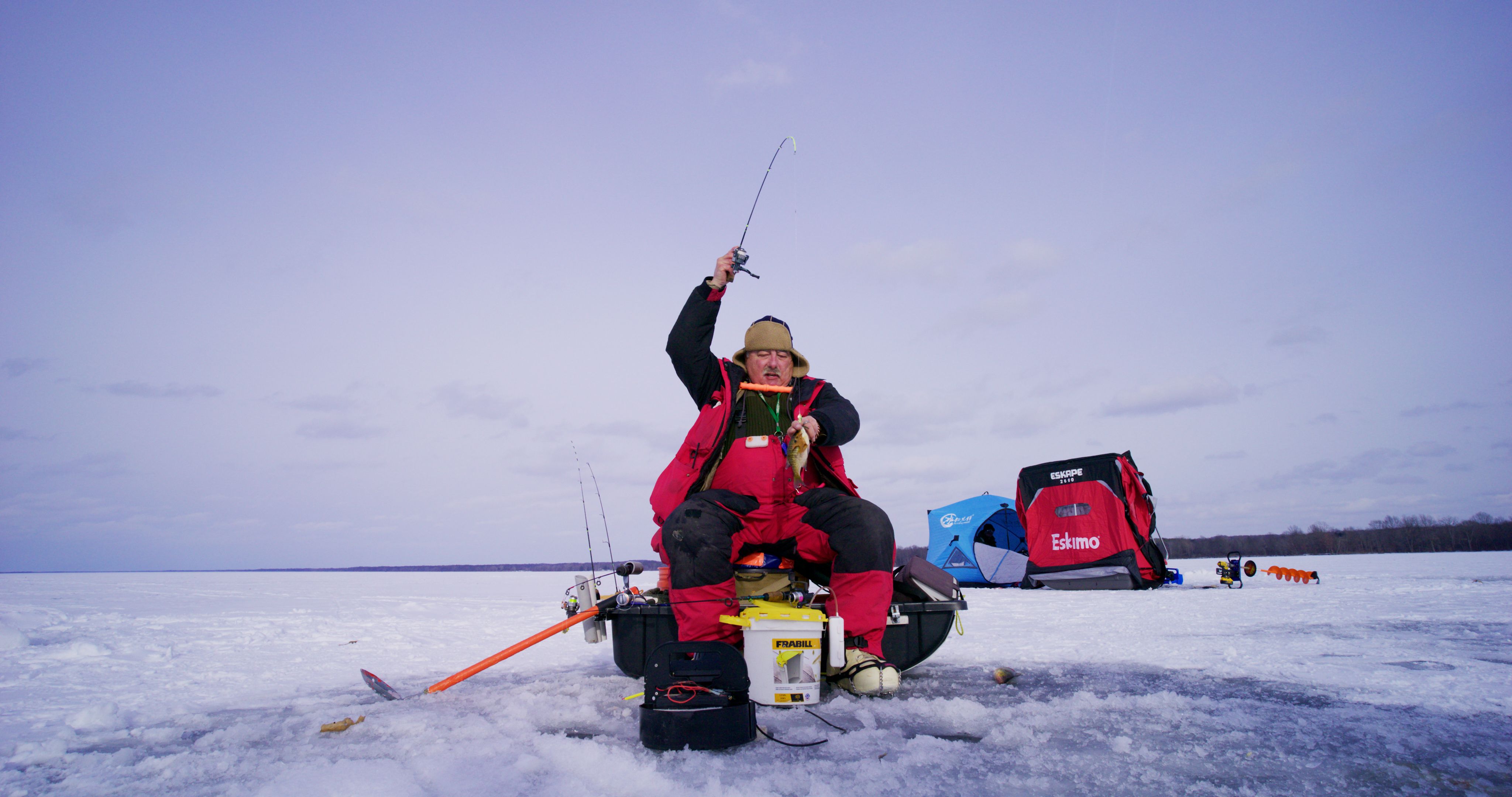 Man in red and black winter gear ice fishing on  afrozen lake, holding a fishing rod with a fish, seated on a crate with ice fishing gear and tents in background.
