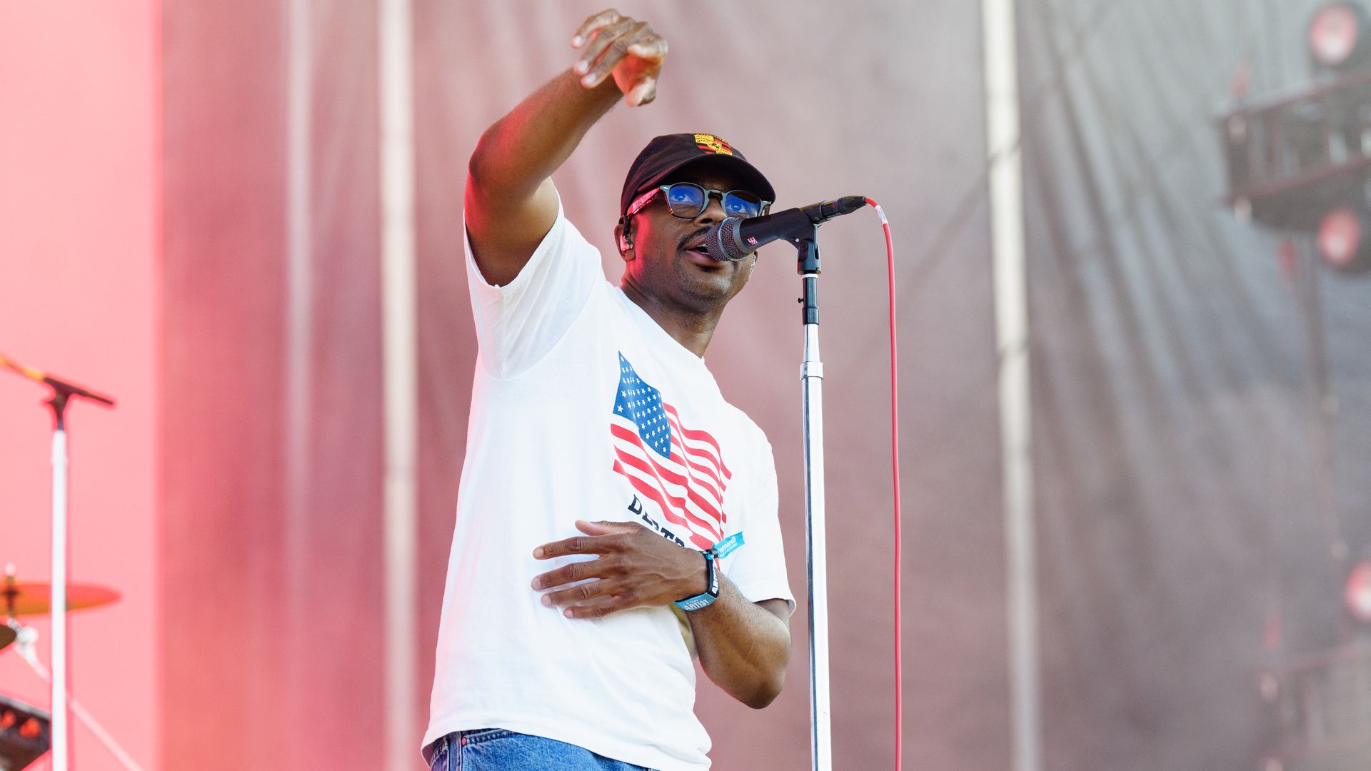A male singer on stage wearing a white T-shirt with an American flag graphic, blue jeans, a black cap, and sunglasses, singing into a microphone with red stage lighting in the background.