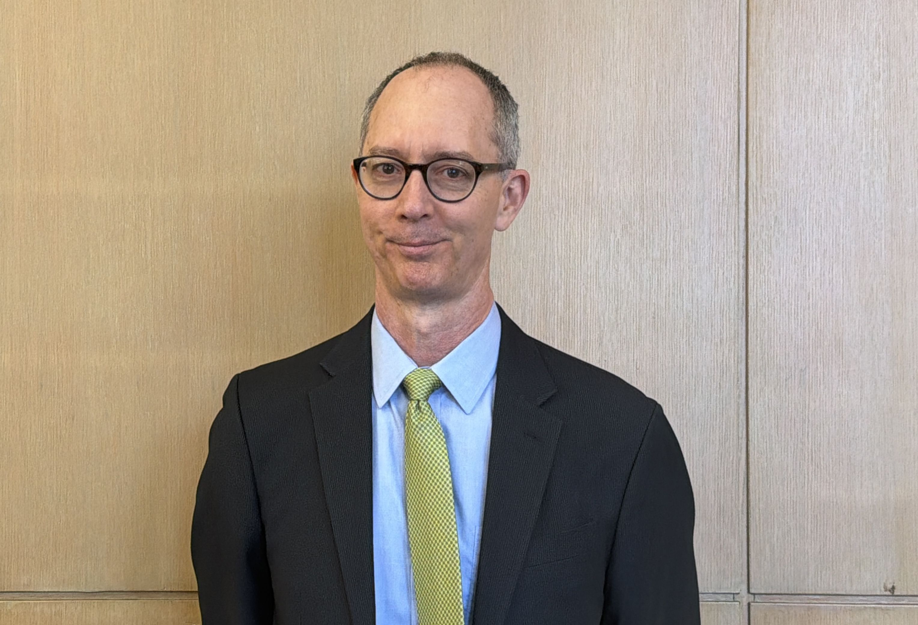 A middle-aged man with short gray hair and glasses, wearing a dark suit, light blue shirt, and yellow tie, stands against a wood-paneled wall.