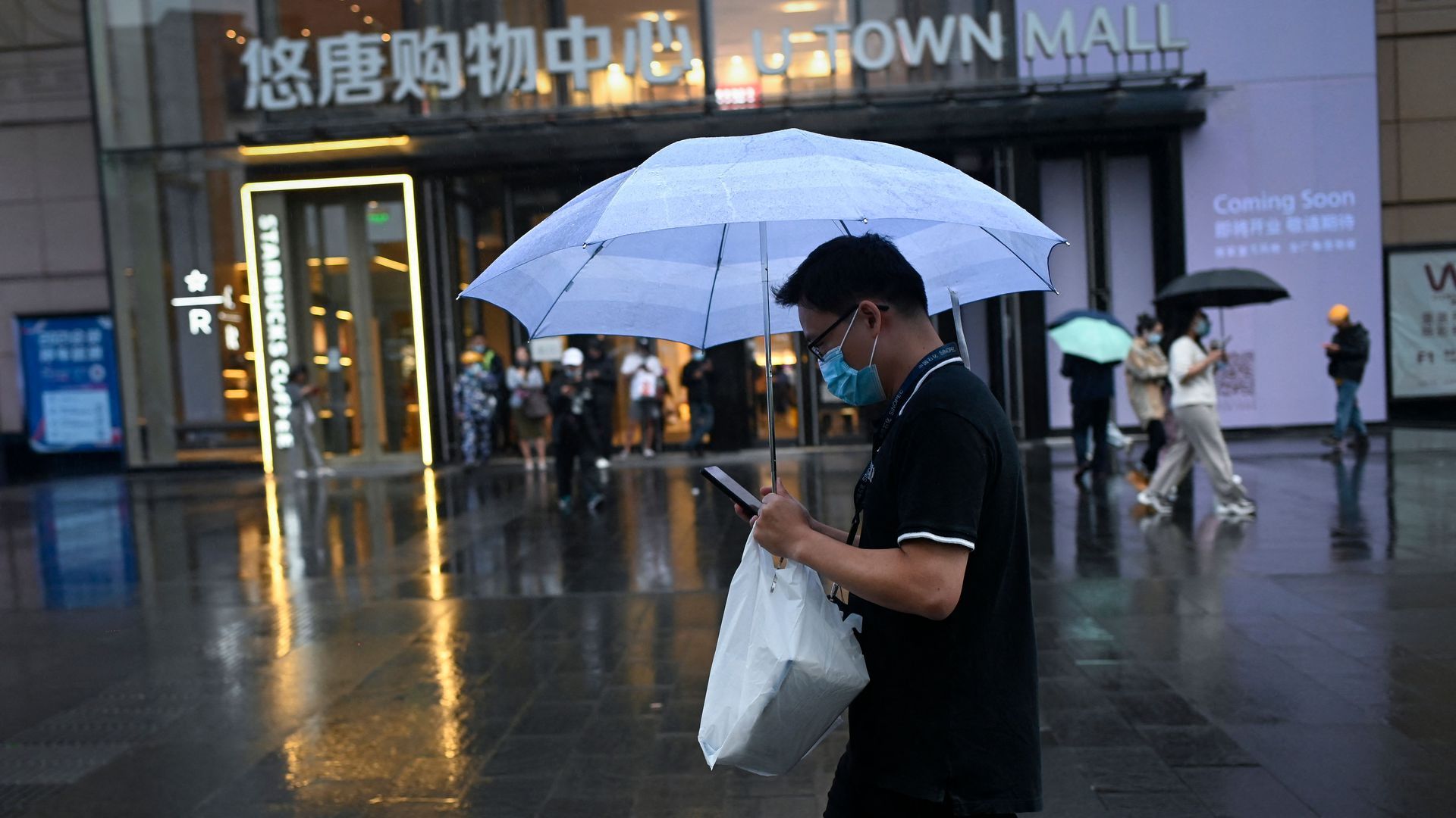 Photo of a masked person holding an umbrella and a plastic bag as they walk past a building