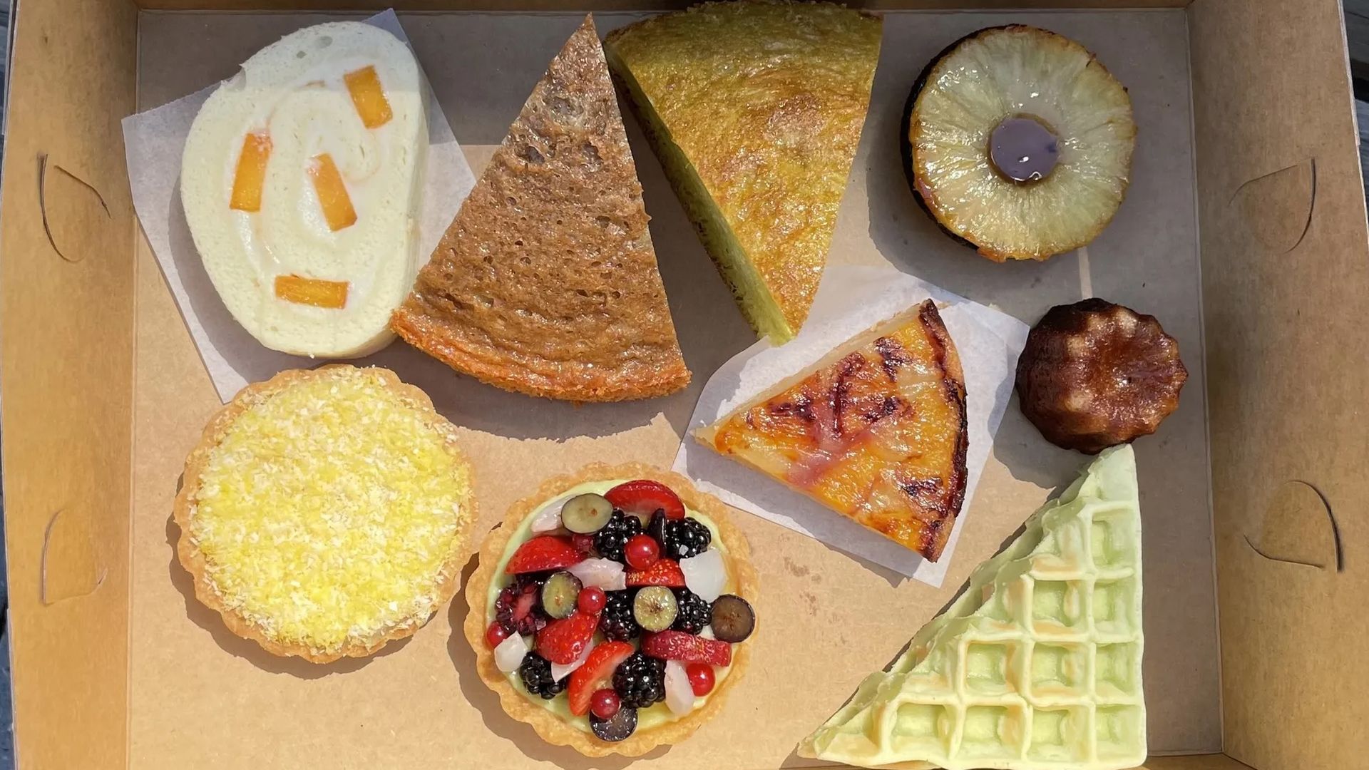 A box of assorted pastries including a fruit tart, lemon tart, rolled cake with candied fruit, waffle slice, various cake slices, a pineapple-topped dessert, and a small canelé, arranged neatly.