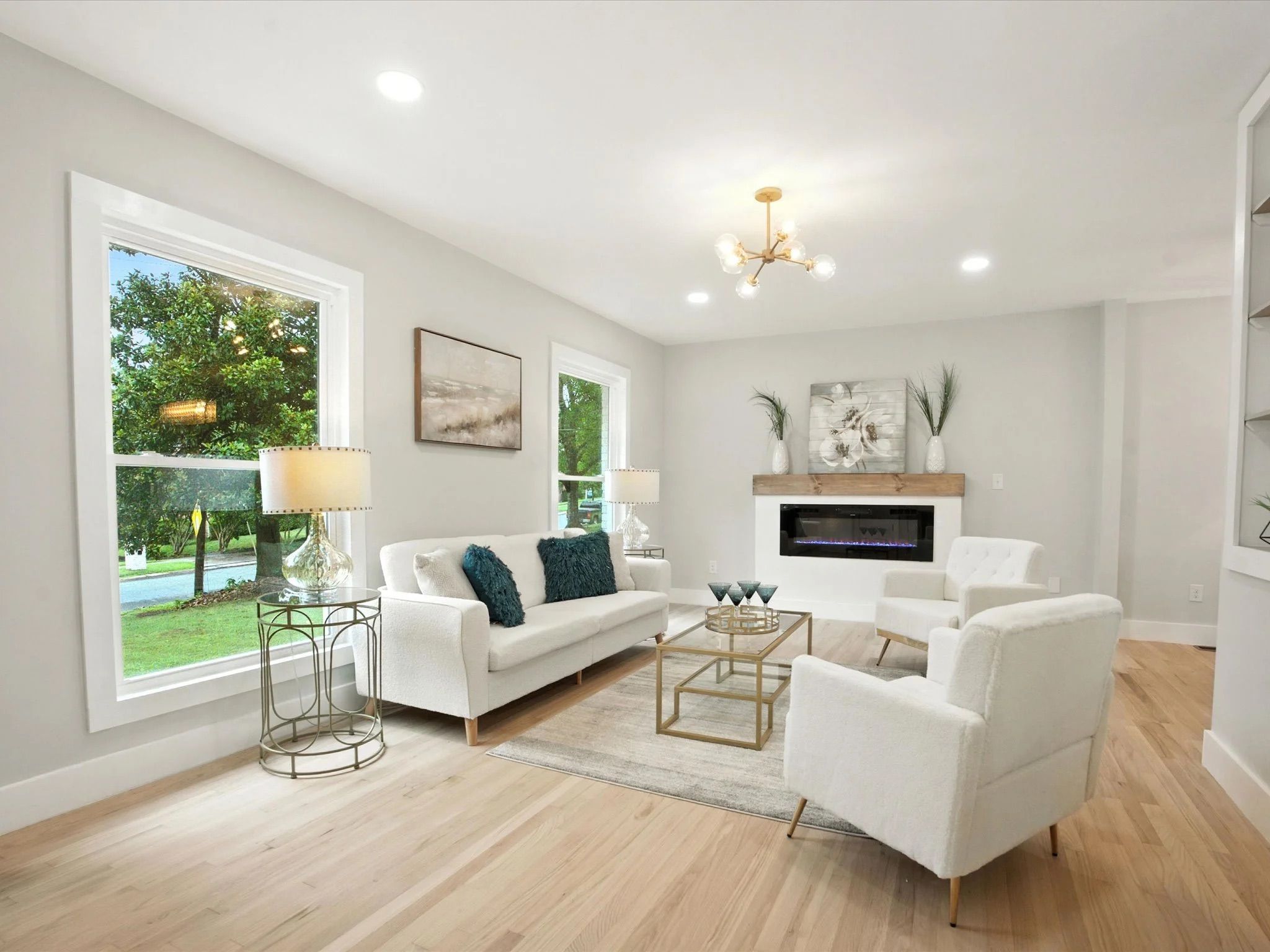 Bright living room with white sofa, two white armchairs, blue cushions, glass coffee table, round metal side table with lamp, electric fireplace, and large windows showing green trees outside.