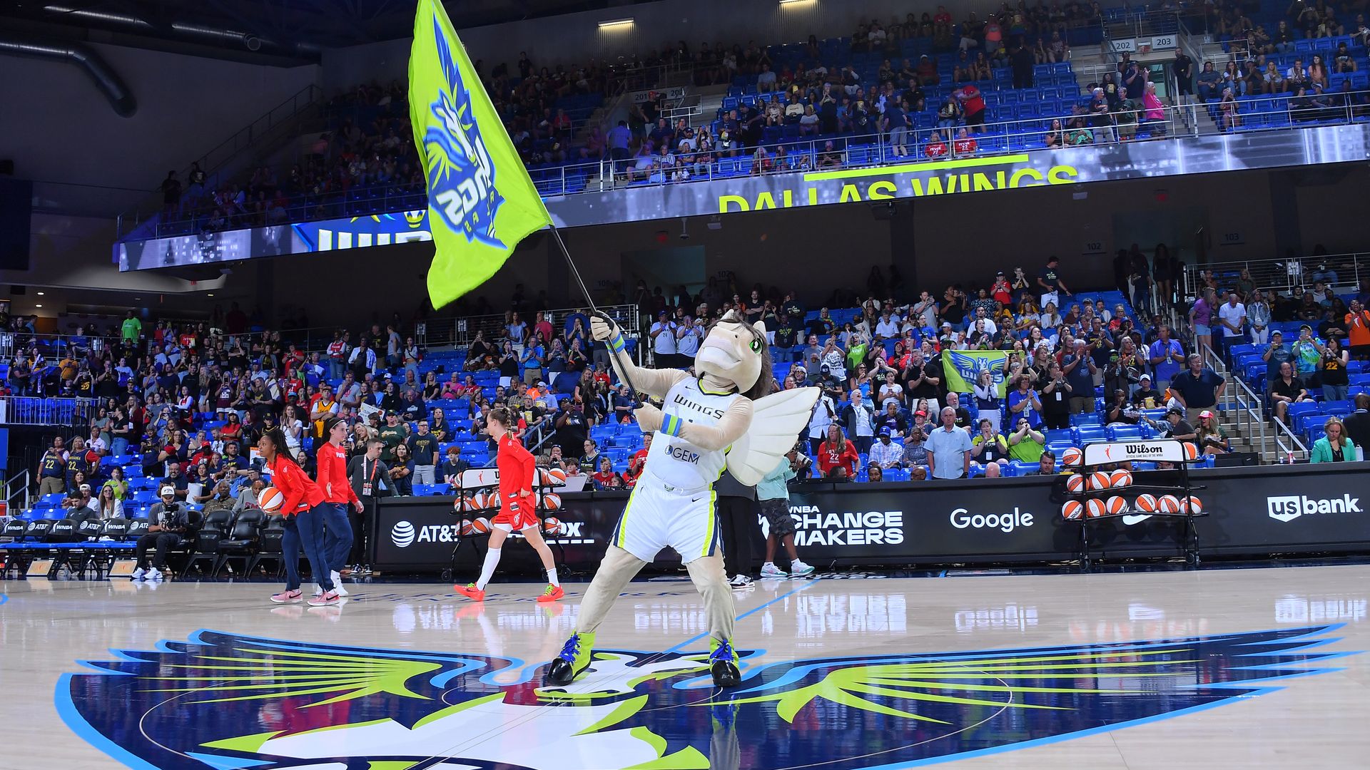 A mascot wearing white waves a green and blue flag in an arena