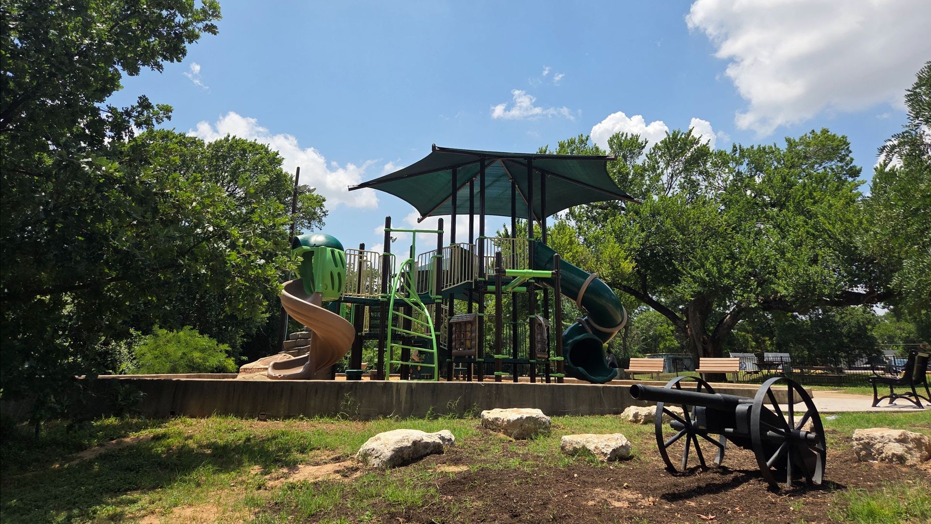 Playground with green and tan slides, climbing structures under a dark green canopy, surrounded by trees, benches, and a small black cannon sculpture on grassy ground with rocks.