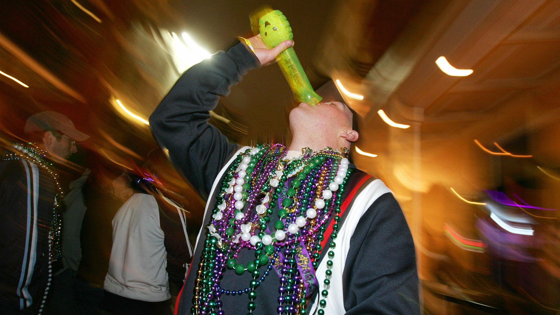 A man wearing Mardi Gras beads leans back to down the last dregs of a drink from a tall, bright green plastic cup with a hand grenade shape on the bottom.