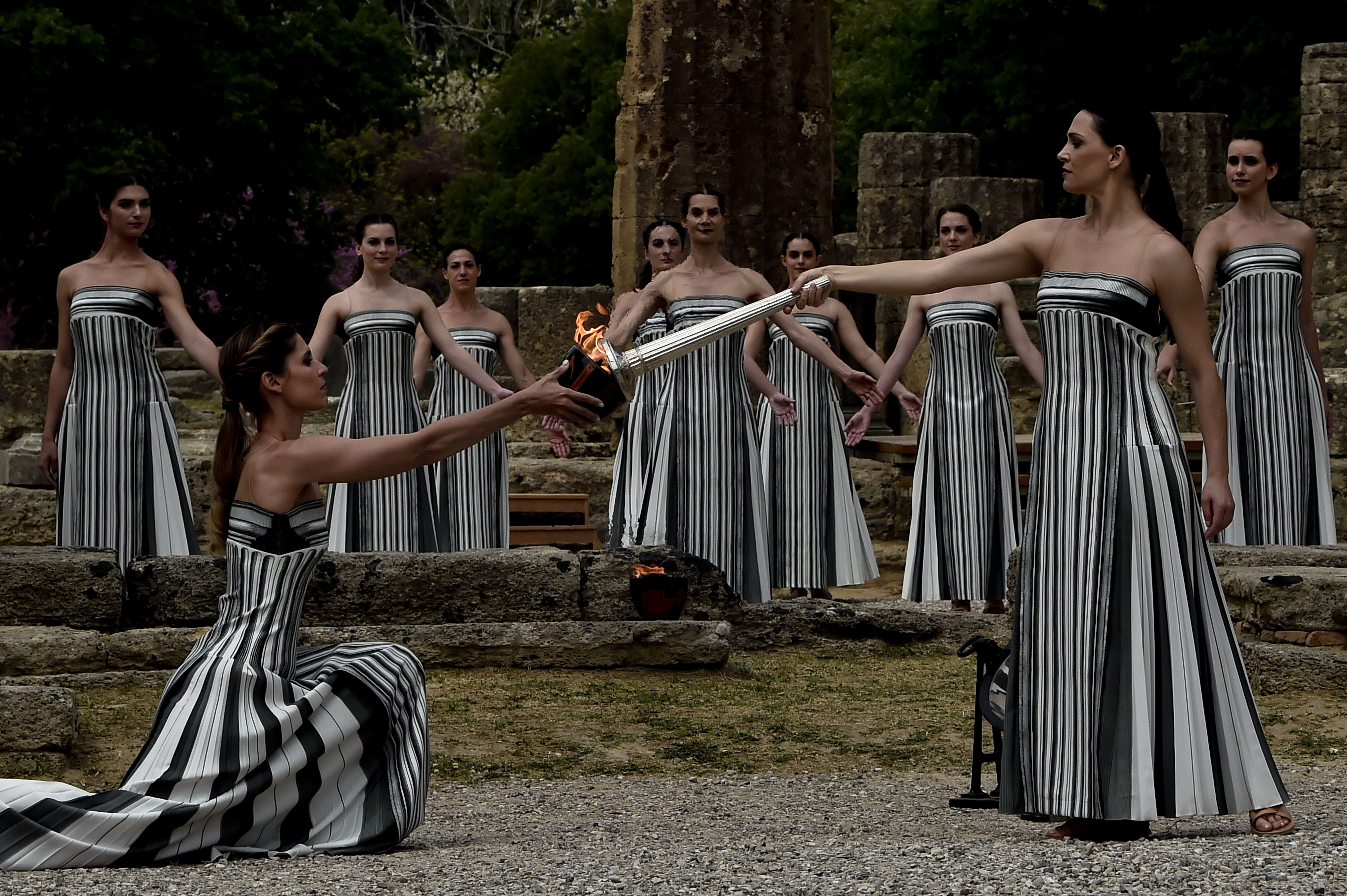 Several women stand in long black and white strapless dresses. In the front, one is kneeling and lighting the torch, held by another who is standing. About nine people are standing in the background with arms outstretched to either side.