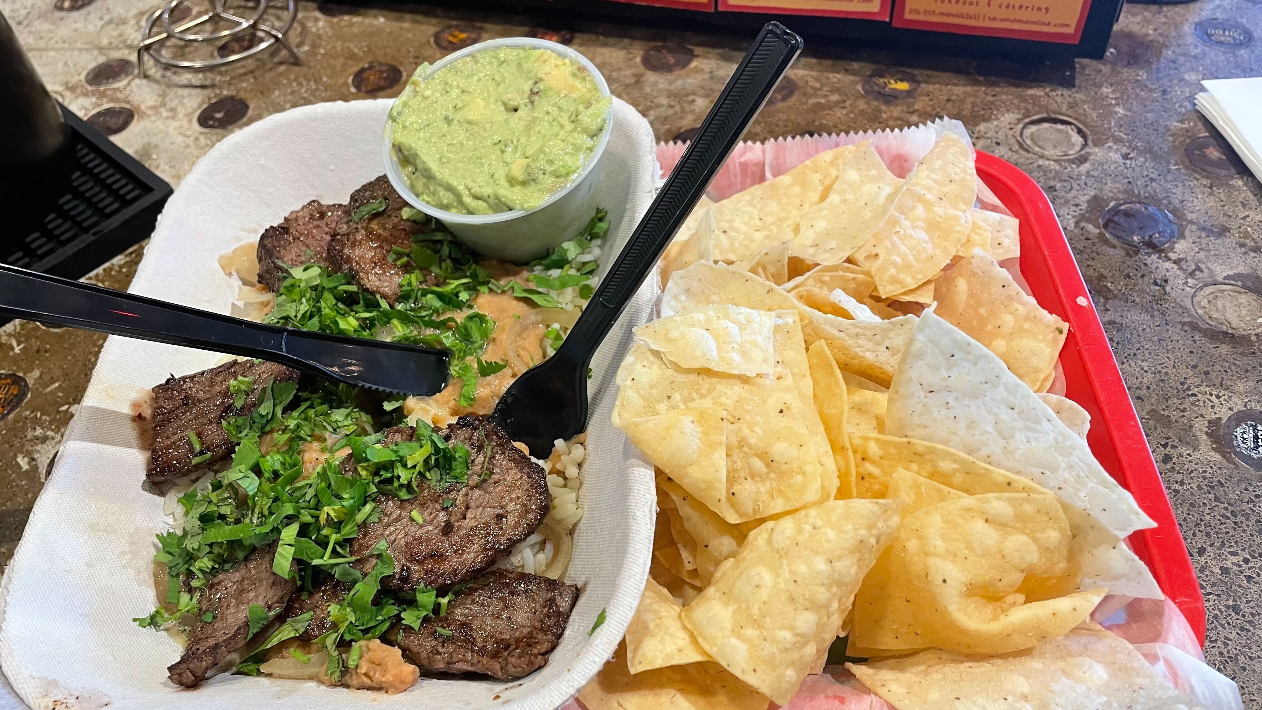 Takeout plate with sliced grilled steak topped with chopped cilantro over rice, a cup of guacamole, and a pile of tortilla chips in a red basket with black plastic utensils.