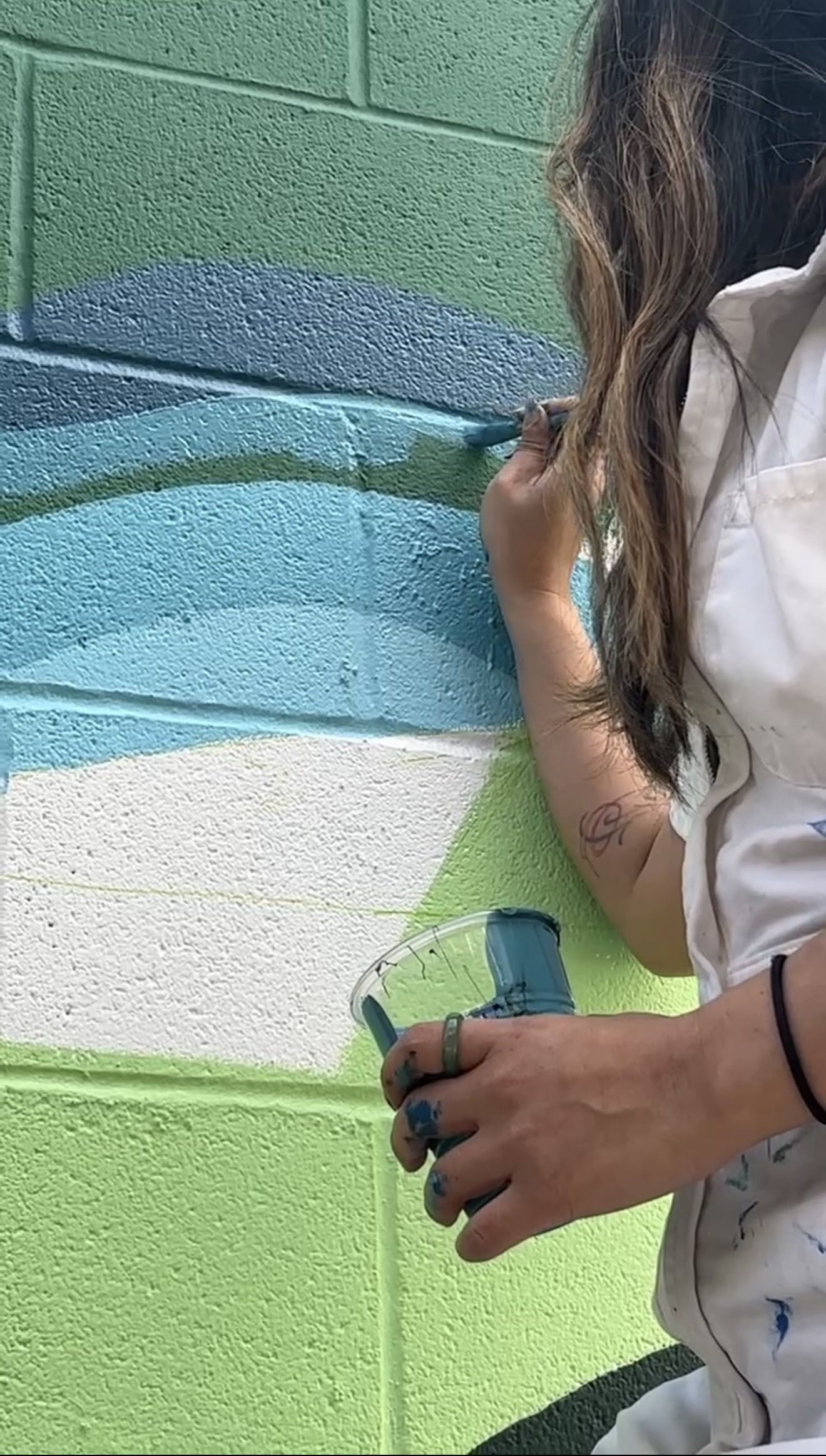 Person with long hair and a white shirt paints a mural on a green and blue cinder block wall, using a teal paint cup to apply blue-green strokes; a small forearm tattoo is visible.