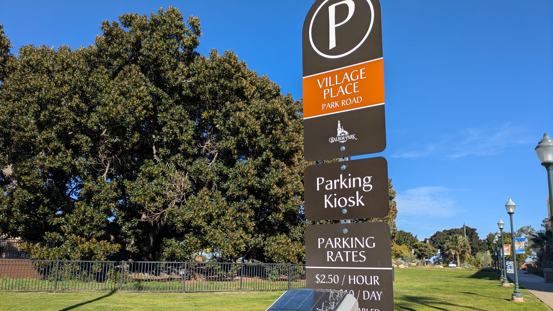 Tall signpost with panels "Village Place Park Road", "Parking Kiosk", and "Parking Rates" beside a gray parking kiosk in a sunny park with large trees and blue sky.