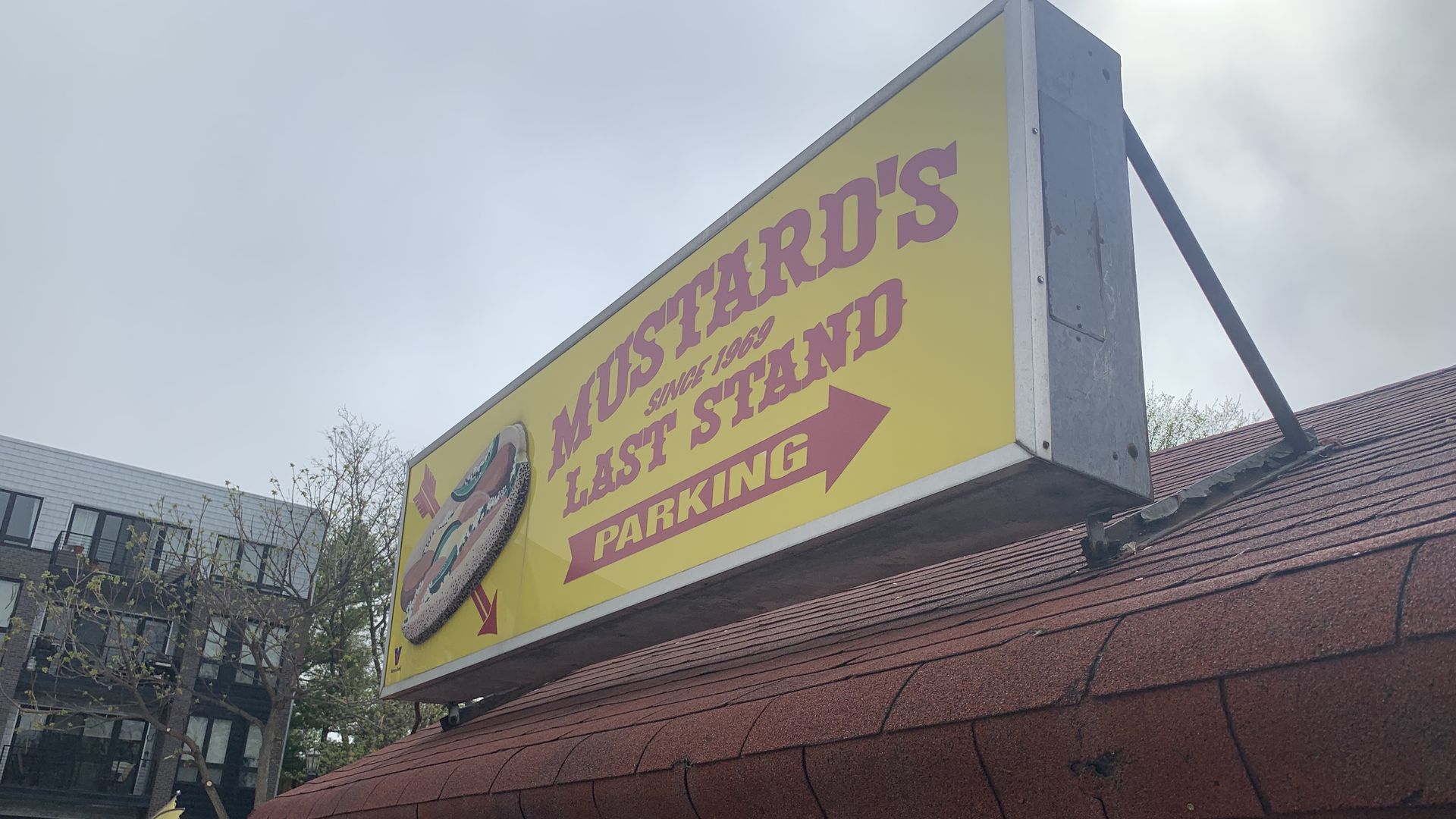 Yellow sign with pink text reading "Mustard's Last Stand" and a right-pointing arrow "Parking"; mounted on a metal frame above a red-tiled roof with a pale blue sky behind.