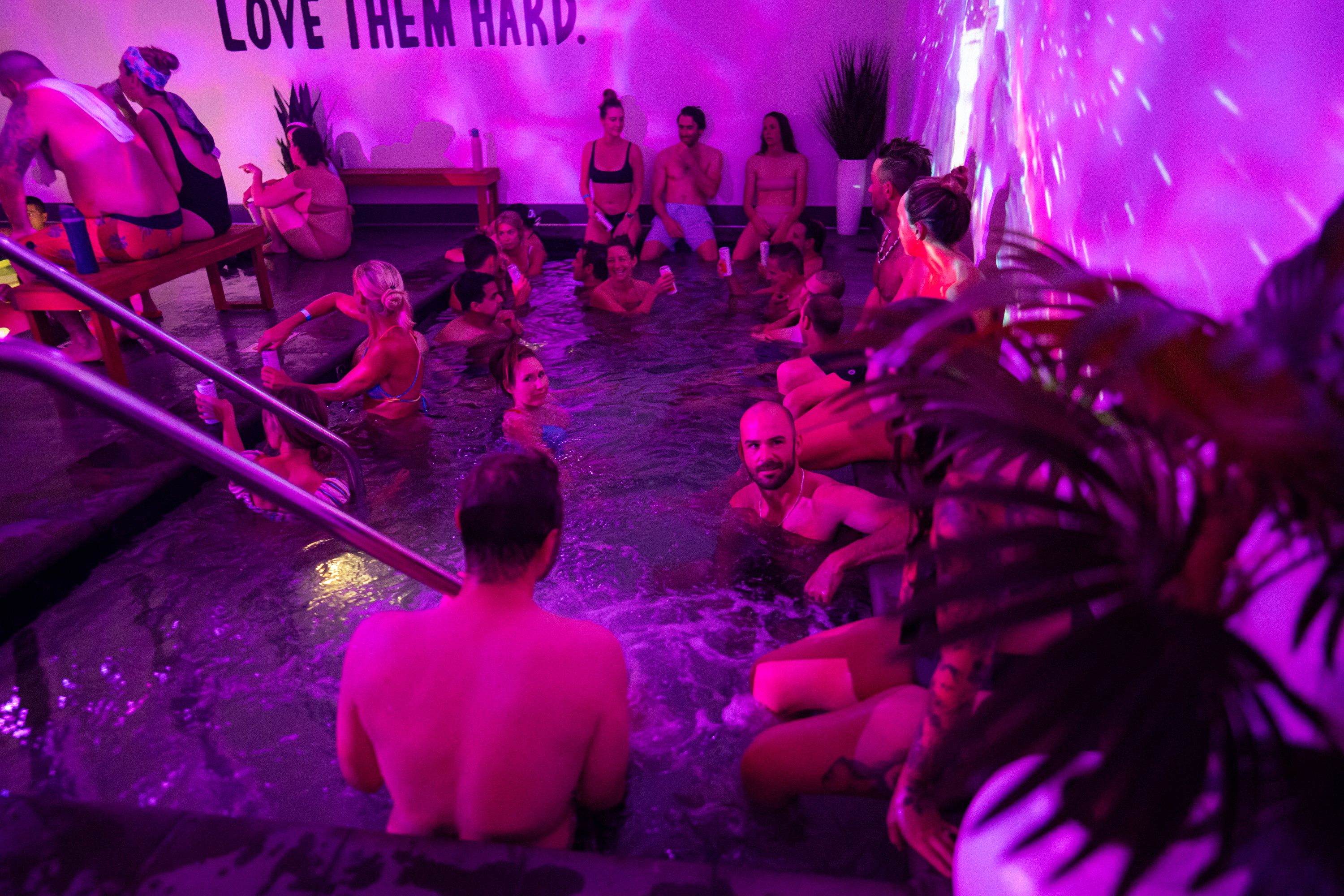 Indoor pool party bathed in purple light; many people in swimsuits sit or stand around a shallow pool, holding drinks, some on benches, as a wall sign reads "LOVE THEM HARD."