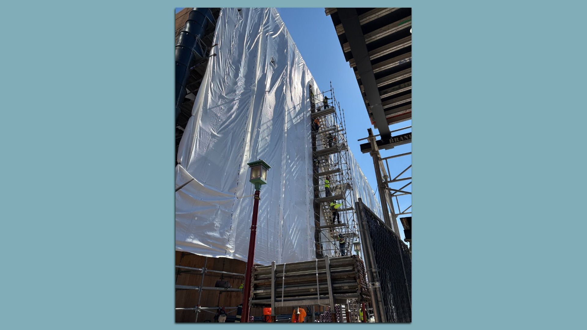 Construction site with a tall white tarp-covered wall, metal scaffolding along the side, workers in yellow safety vests climbing, blue sky visible, chain-link fence and steel beams nearby.