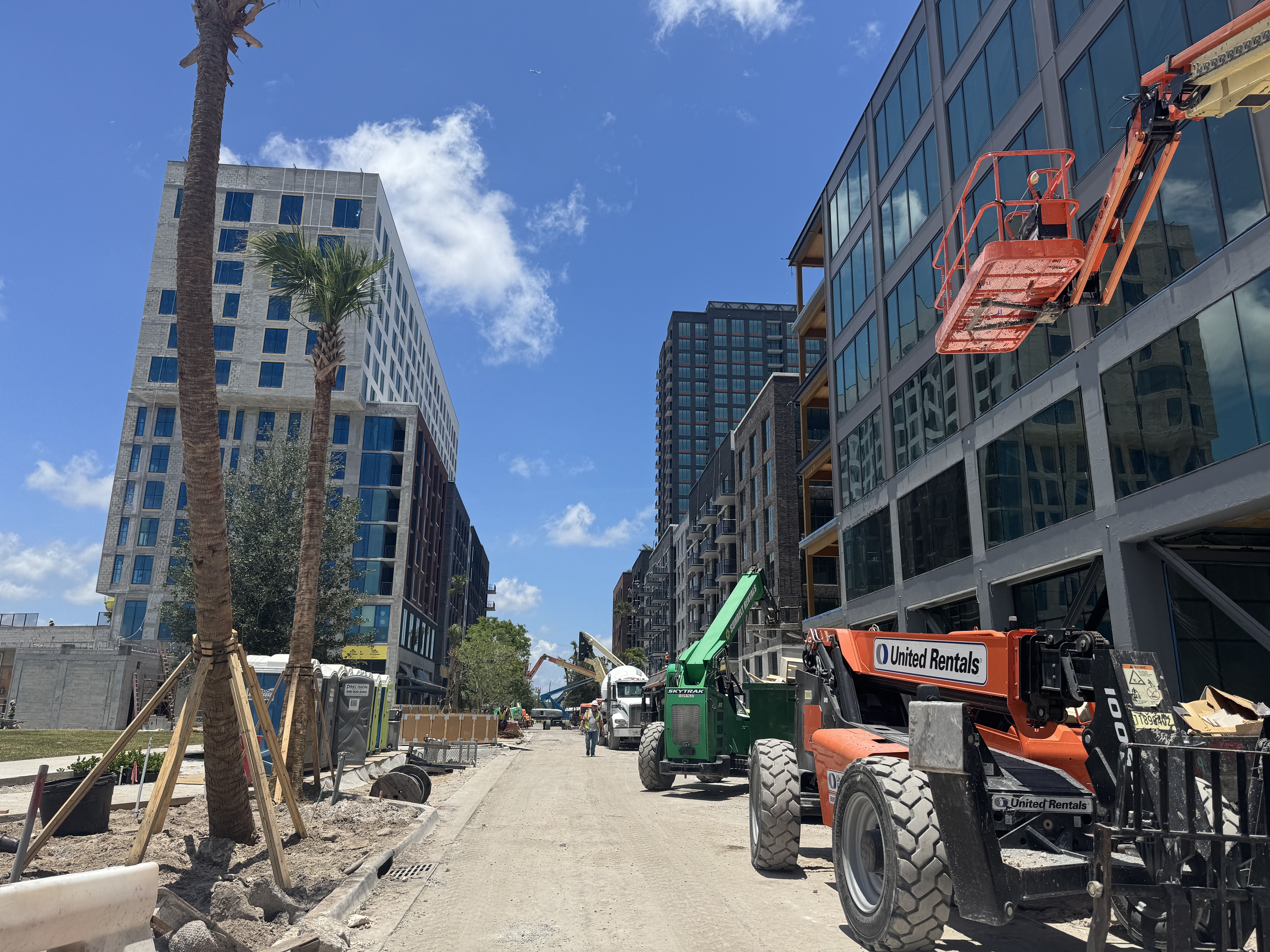 Sunny city construction scene with a glass-front building under construction on the right and tall palm trees on the left. An orange aerial lift and other construction equipment occupy the street.