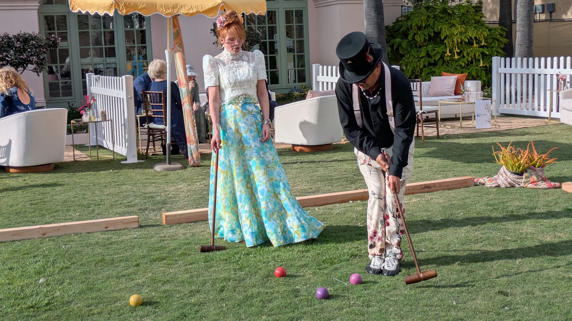 Two people in vintage attire croquet on a sunny lawn: a woman in white lace and a blue floral skirt under a yellow parasol, and a man in a top hat; pastel building and white fence in the background.