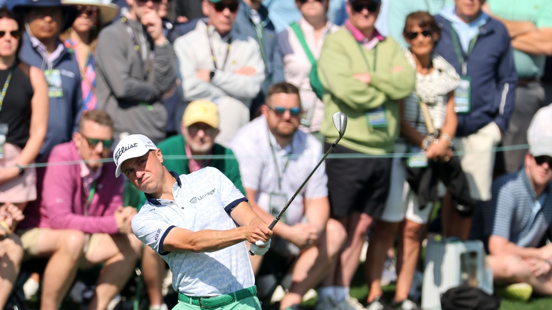 A person in a white polo shirt swings a golf club while a crowd watches