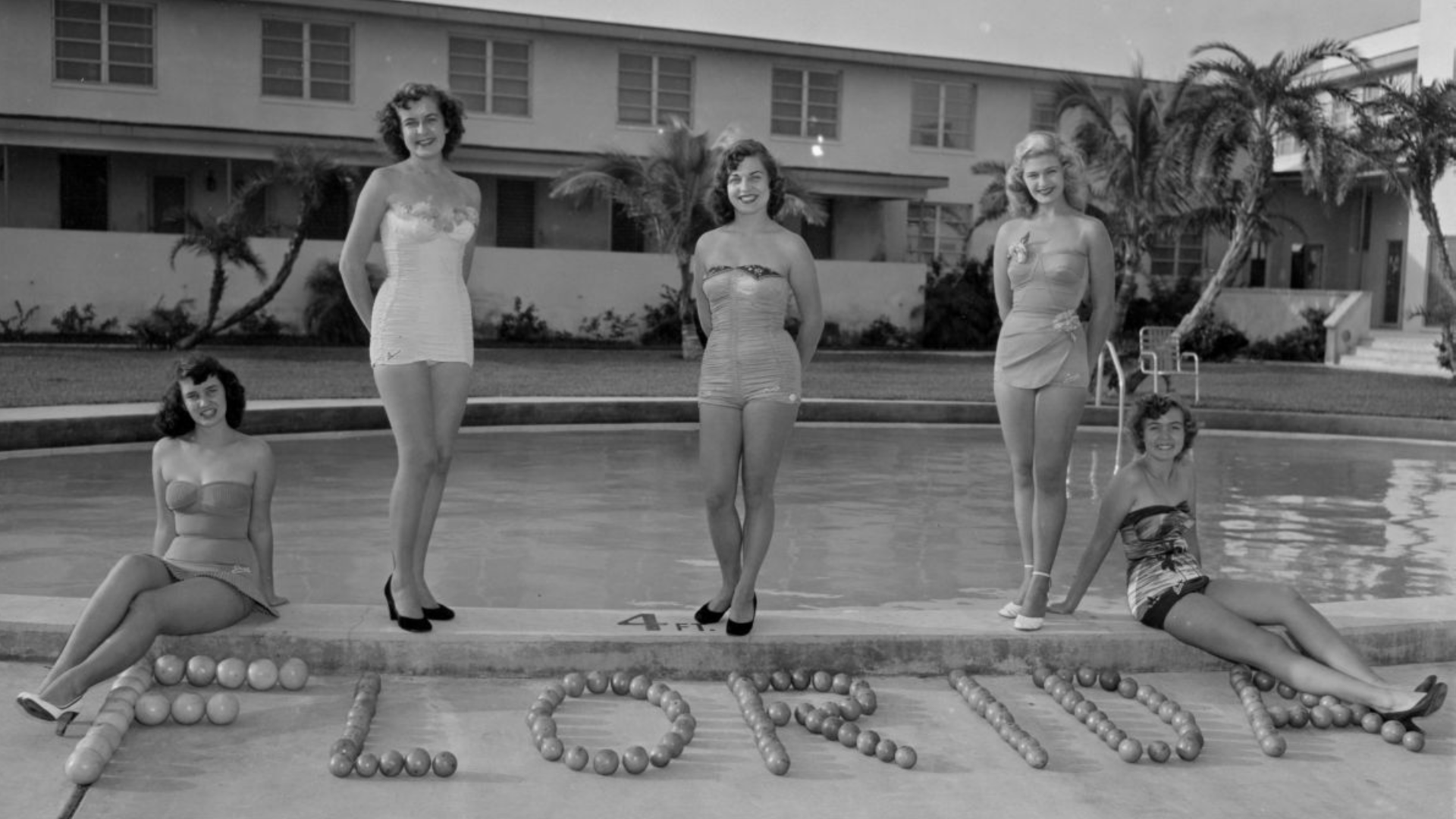 Four women in vintage bathing suits pose in front of a "Florida" spelled out in citrus.