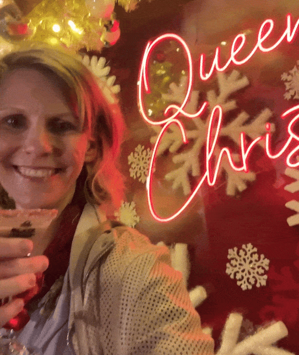 Gif of woman cheersing a martini glass in front of a neon sign that says "Queen of Christmas."