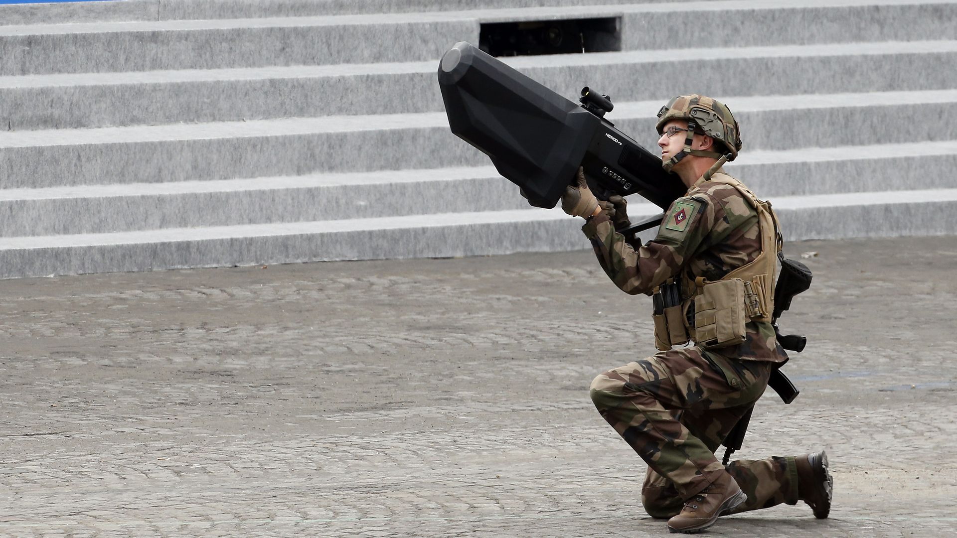 A man crouches in military fatigues and points a rifle-like device toward the sky
