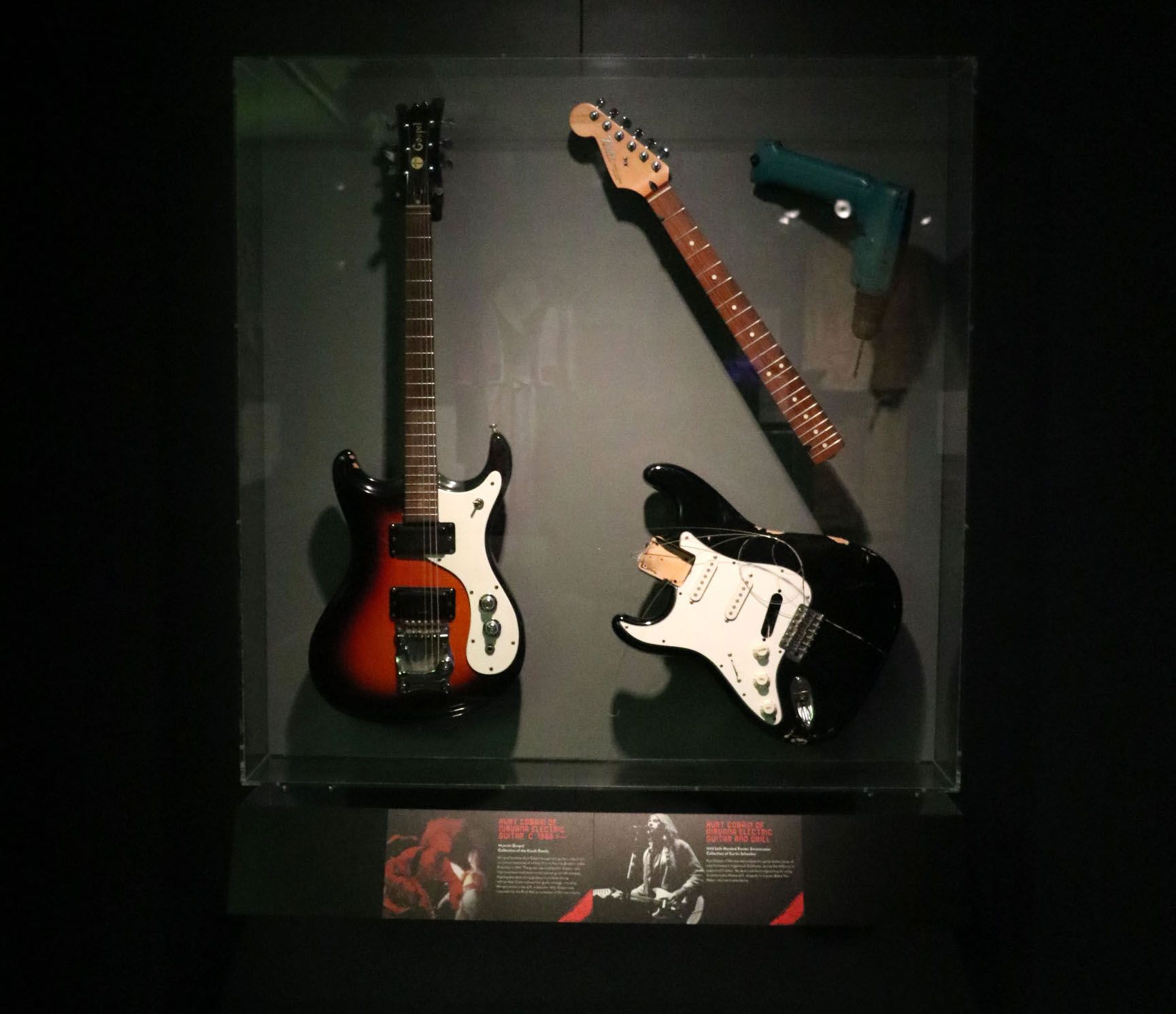 Two Kurt Cobain electric guitars in a case against a wall, one with the fret snapped. 