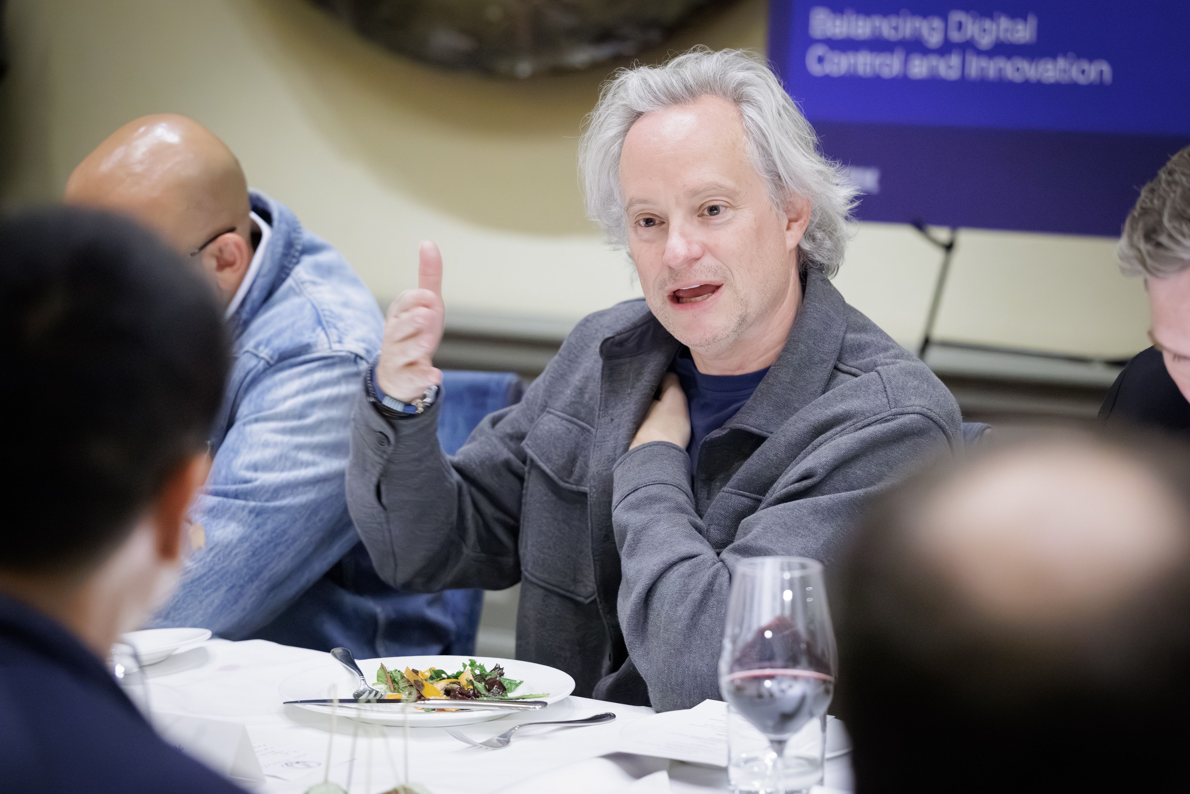 Gray-haired man in a gray jacket speaks at a dinner table, raising his index finger; a plate of salad and a wine glass are in the foreground, with a purple banner in the background.