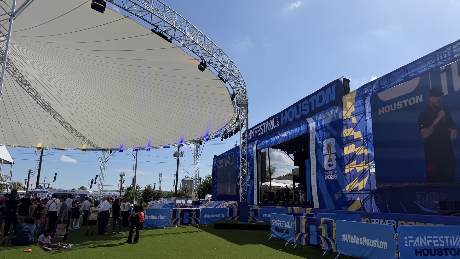 Outdoor festival stage with a white curved fabric canopy supported by metal trusses, blue banners reading HOUSTON and FIFA FAN FESTIVAL, and a crowd standing on green grass under a bright blue sky.
