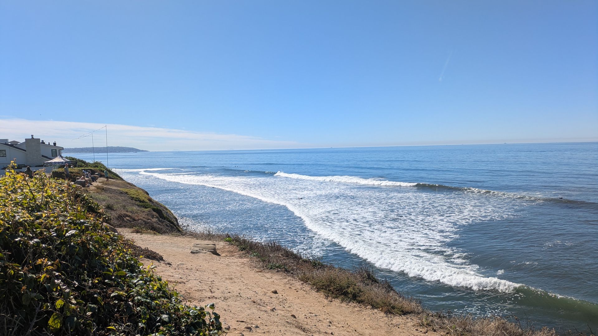 Sunny coastal scene with blue sky, crashing white waves, sandy cliff path, green bushes, and houses with people near the edge overlooking the ocean.