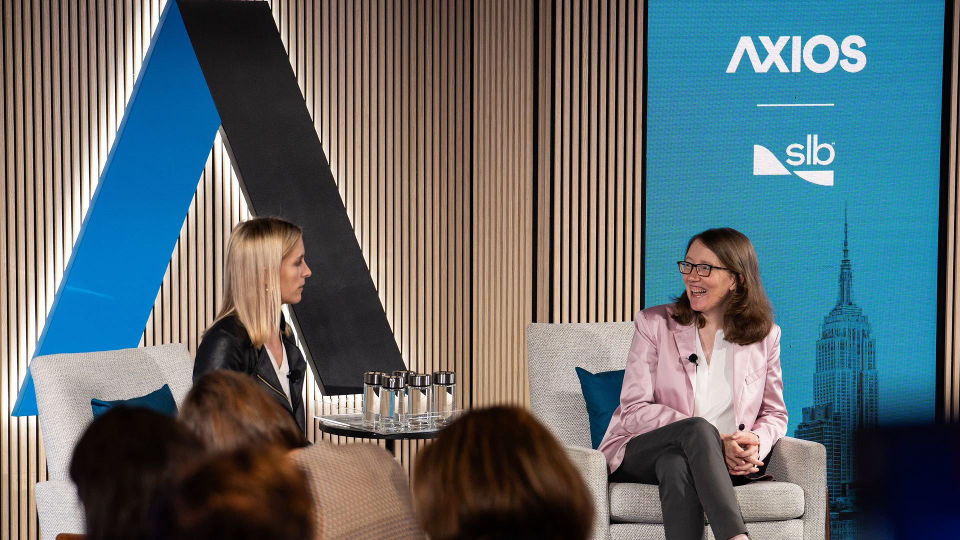 Two women seated in armchairs on a stage, one in a pink blazer smiling, the other wearing a black jacket listening. Audience in foreground, blue and black decor with Axios and SLB logos in background.