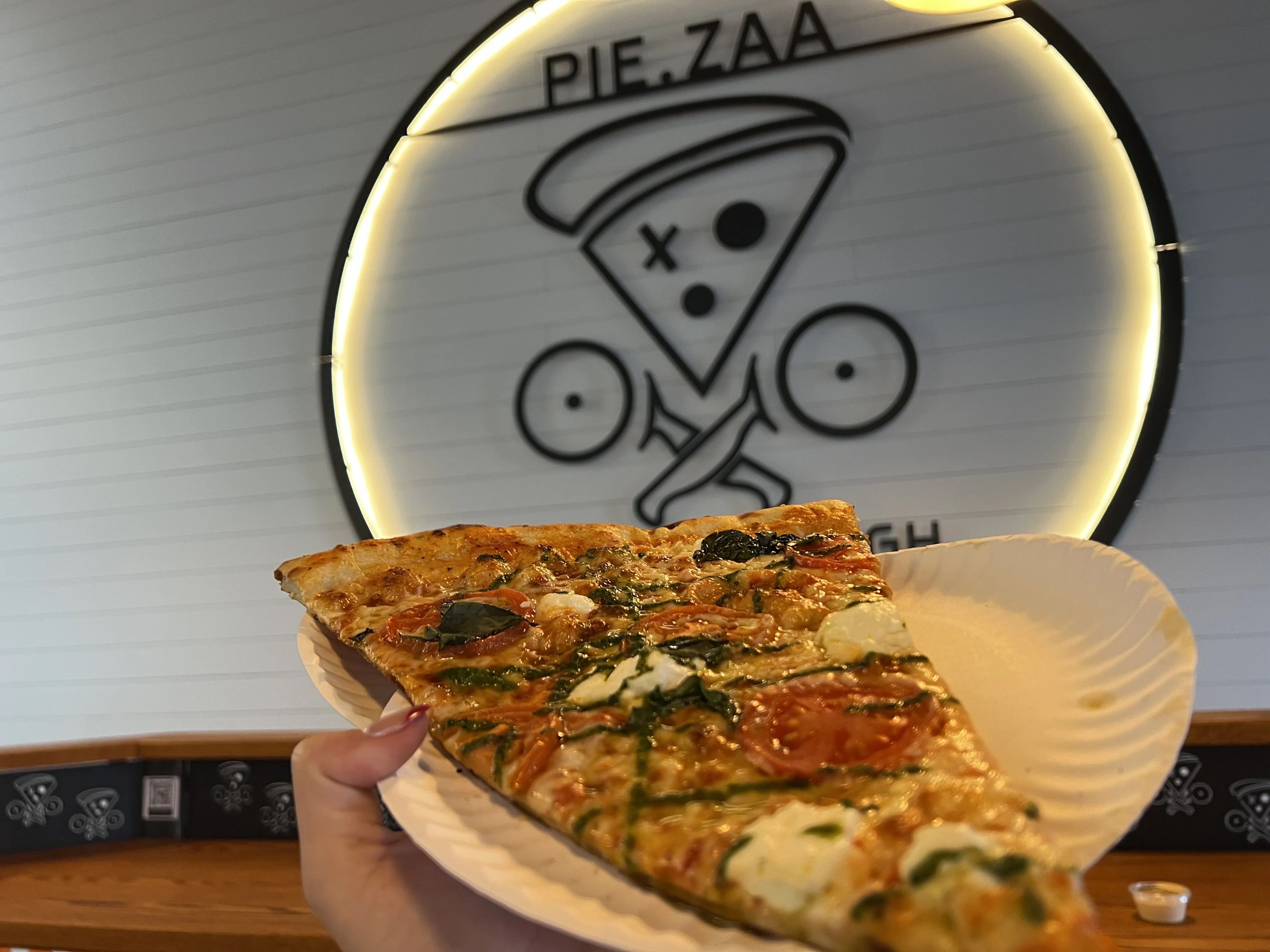 Hand holding a slice of pizza with tomatoes, spinach, and cheese on a white paper plate in front of a circular lit sign featuring a pizza slice and text "PIE.ZAA"
