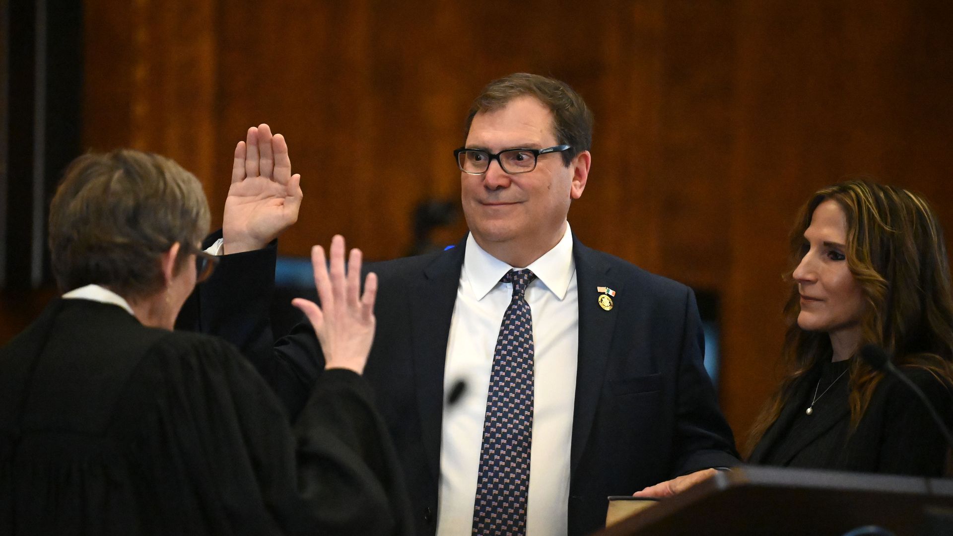 Sarcone holding his hand up as he's sworn in by a judge, wearing glasses, a navy blue suit jacket, white collared shirt and an American flag tie